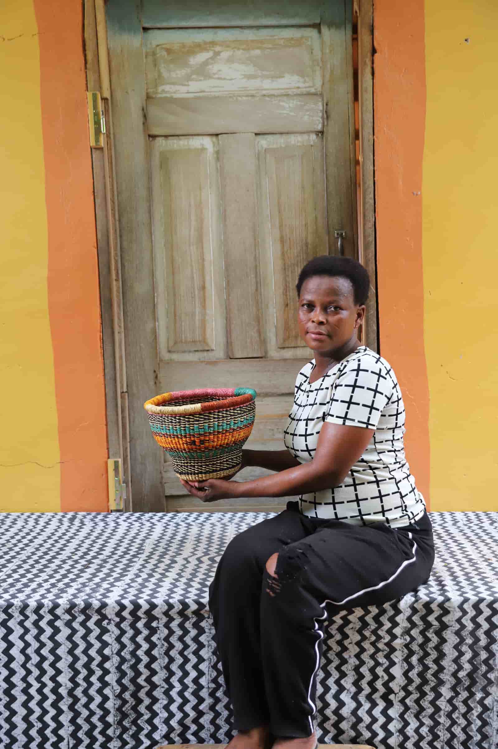 Woman holding a tiny handwoven drum storage basket with teal, amber and dark red stripes, before a wooden door.