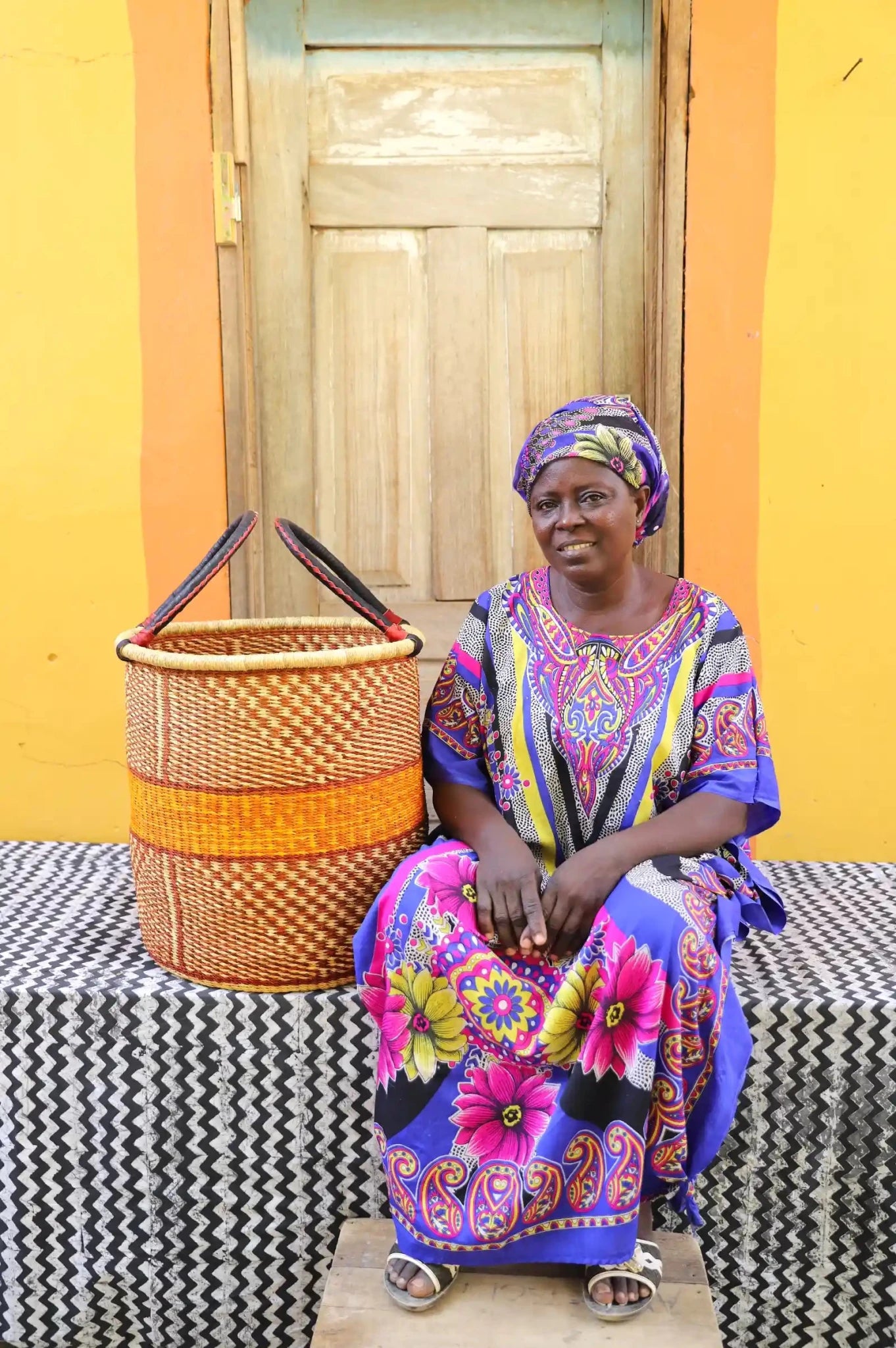 Weaver sitting with colourful hand woven laundry basket on a black and white cloth in front of wooden door
