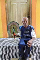 Man holding a handwoven Bolga fan with a green and pink concentric arch pattern, seated before a wooden door.