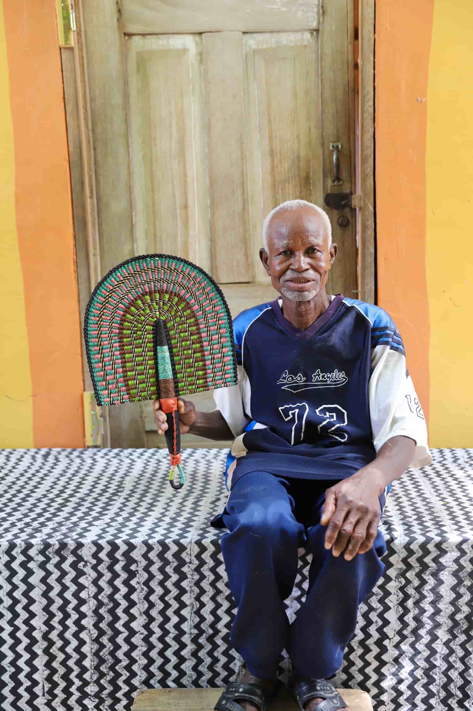 Man holding a handwoven Bolga fan with a green and pink concentric arch pattern, seated before a wooden door.