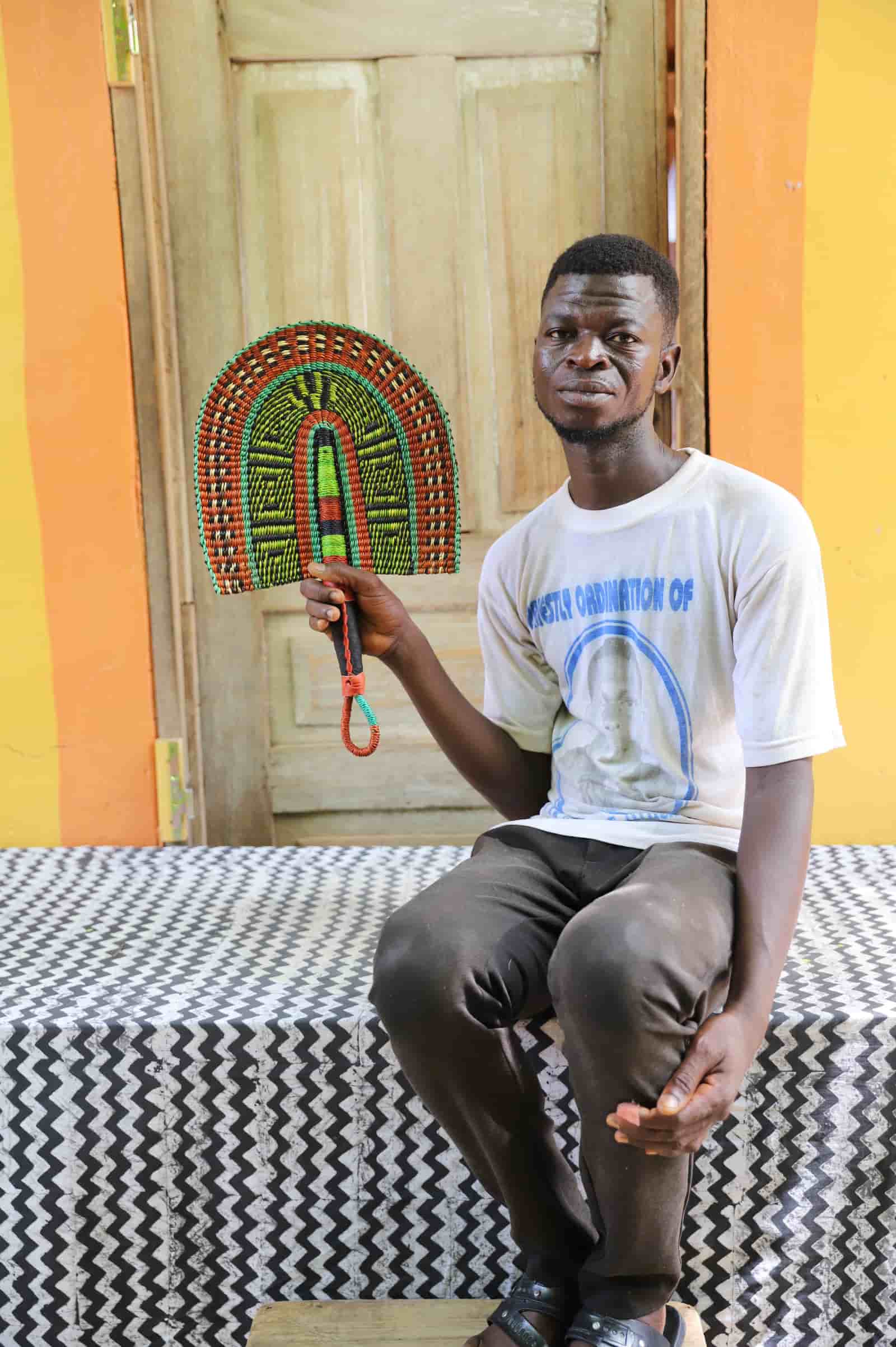 Man holding a handwoven Bolga fan with an orange and lime geometric arch pattern, seated before a wooden door.