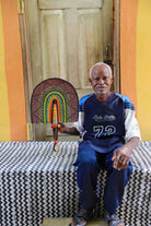 Man holding a handwoven Bolga fan with a multicolour arch pattern in pink, green and amber, seated before a wooden door.