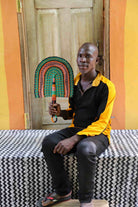 Man seated holding a Bolga fan in green and terracotta concentric arch pattern, before a wooden door with yellow wall.
