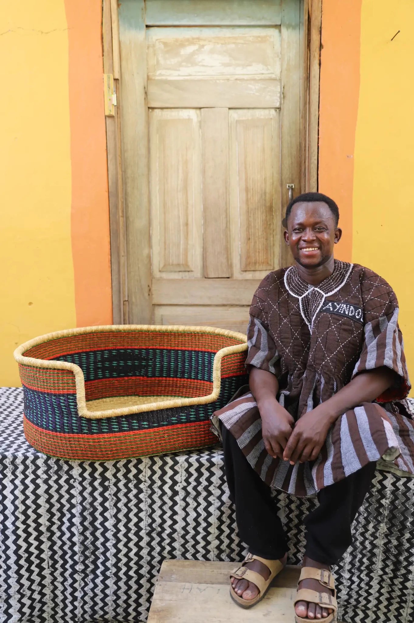 Man sitting on a bench with a woven basket next to a wooden door and yellow wall.