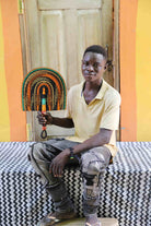 Young man seated holding a Bolga fan in dark brown with amber and teal arch pattern, before a wooden door with yellow wall.