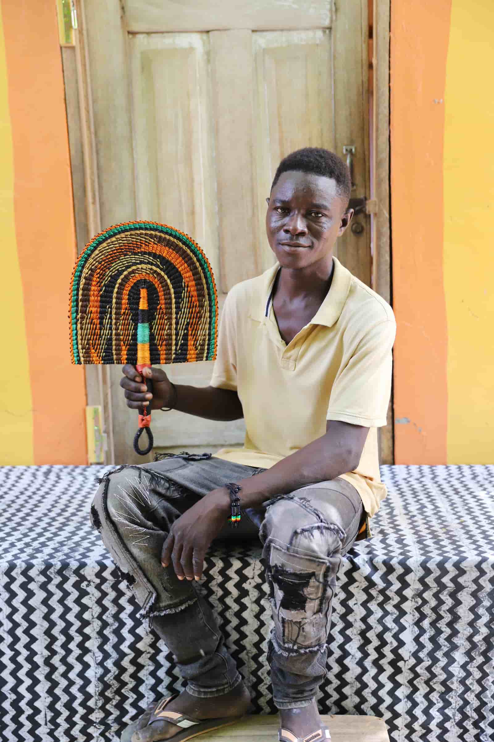 Young man seated holding a Bolga fan in dark brown with amber and teal arch pattern, before a wooden door with yellow wall.