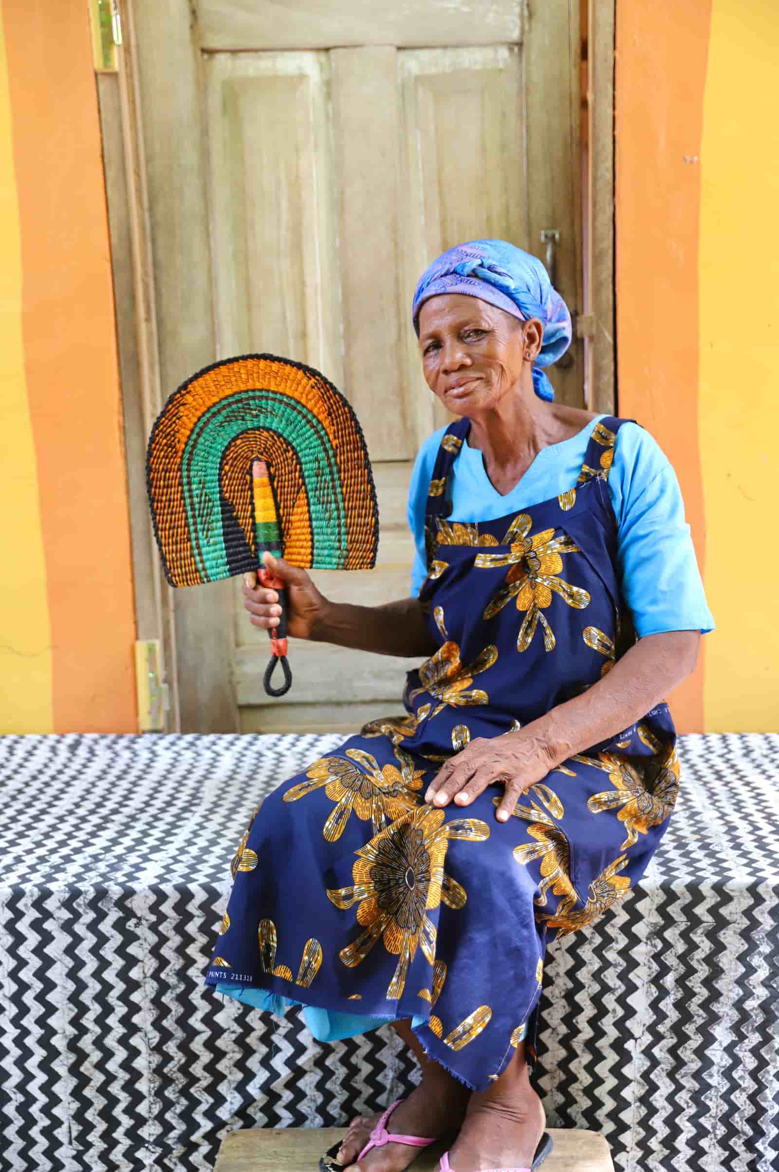 Woman seated holding a Bolga fan in amber and teal concentric arch pattern, before a wooden door with yellow wall.