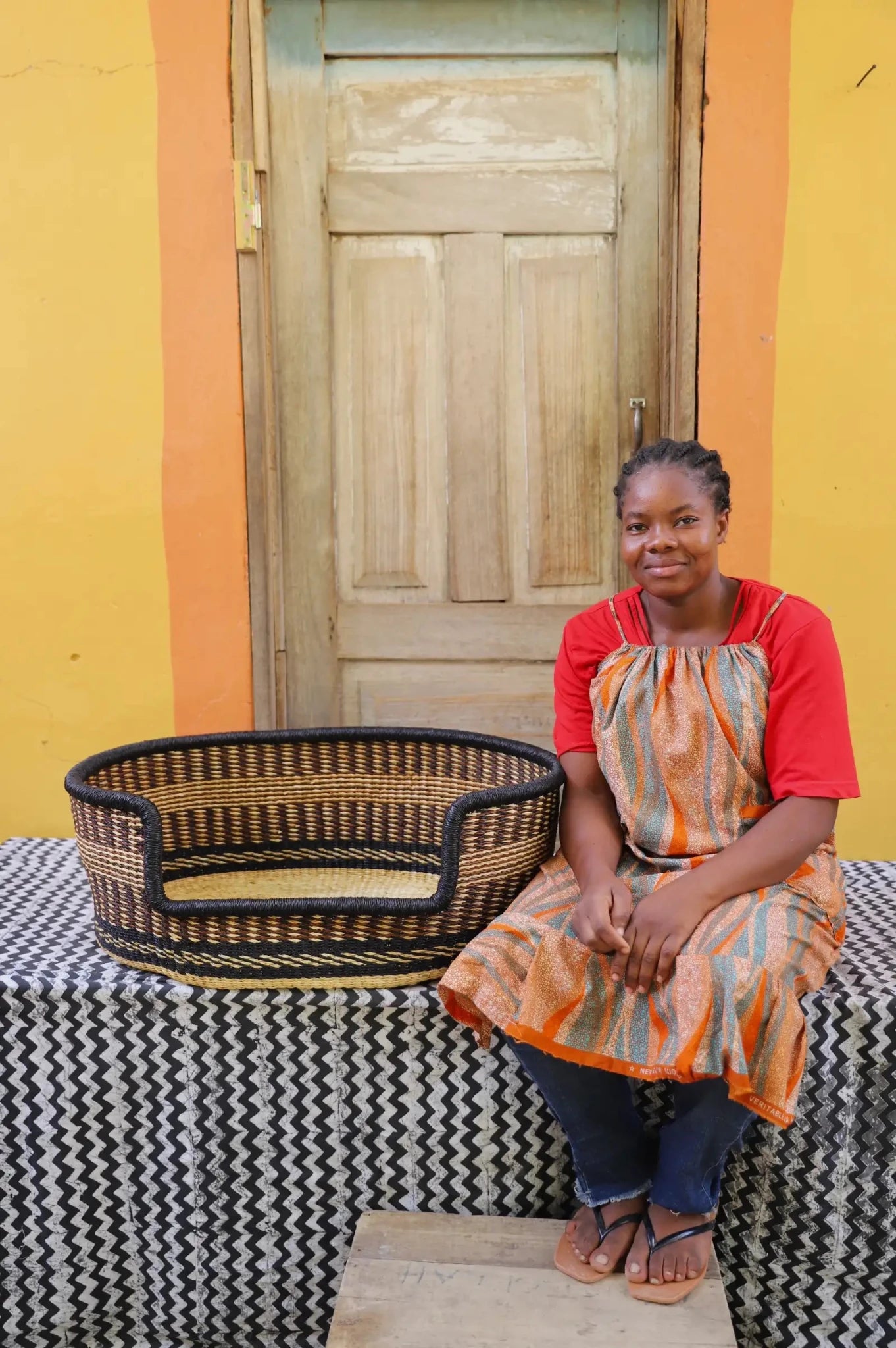 Woman sitting on a bench with a woven basket next to a wooden door and yellow wall.
