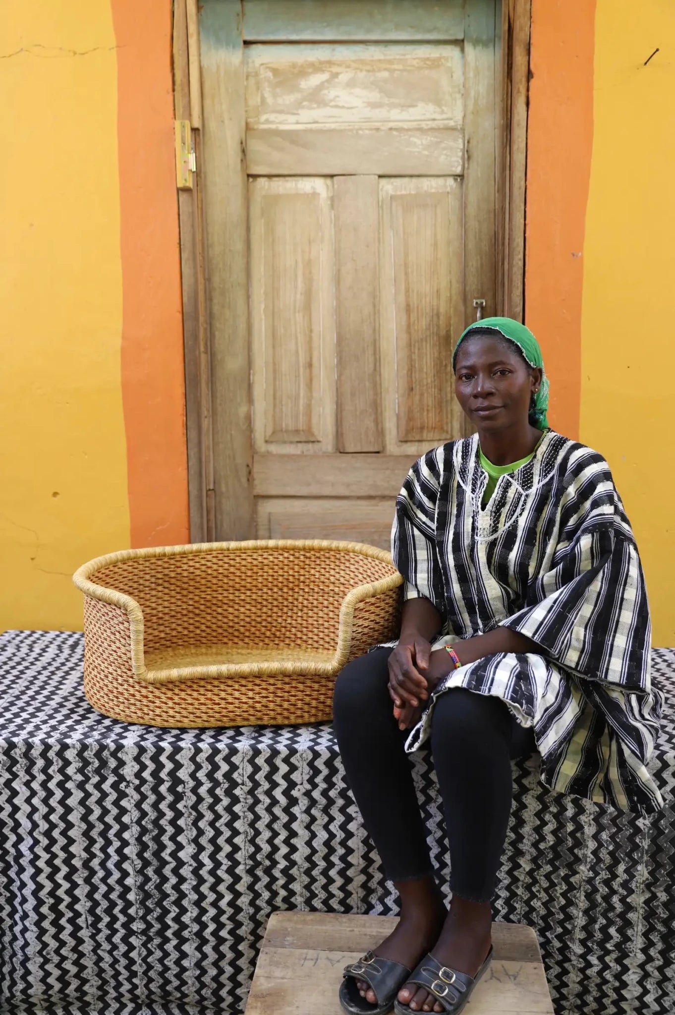 Woman sitting on a bench with a woven basket next to a wooden door and yellow wall.