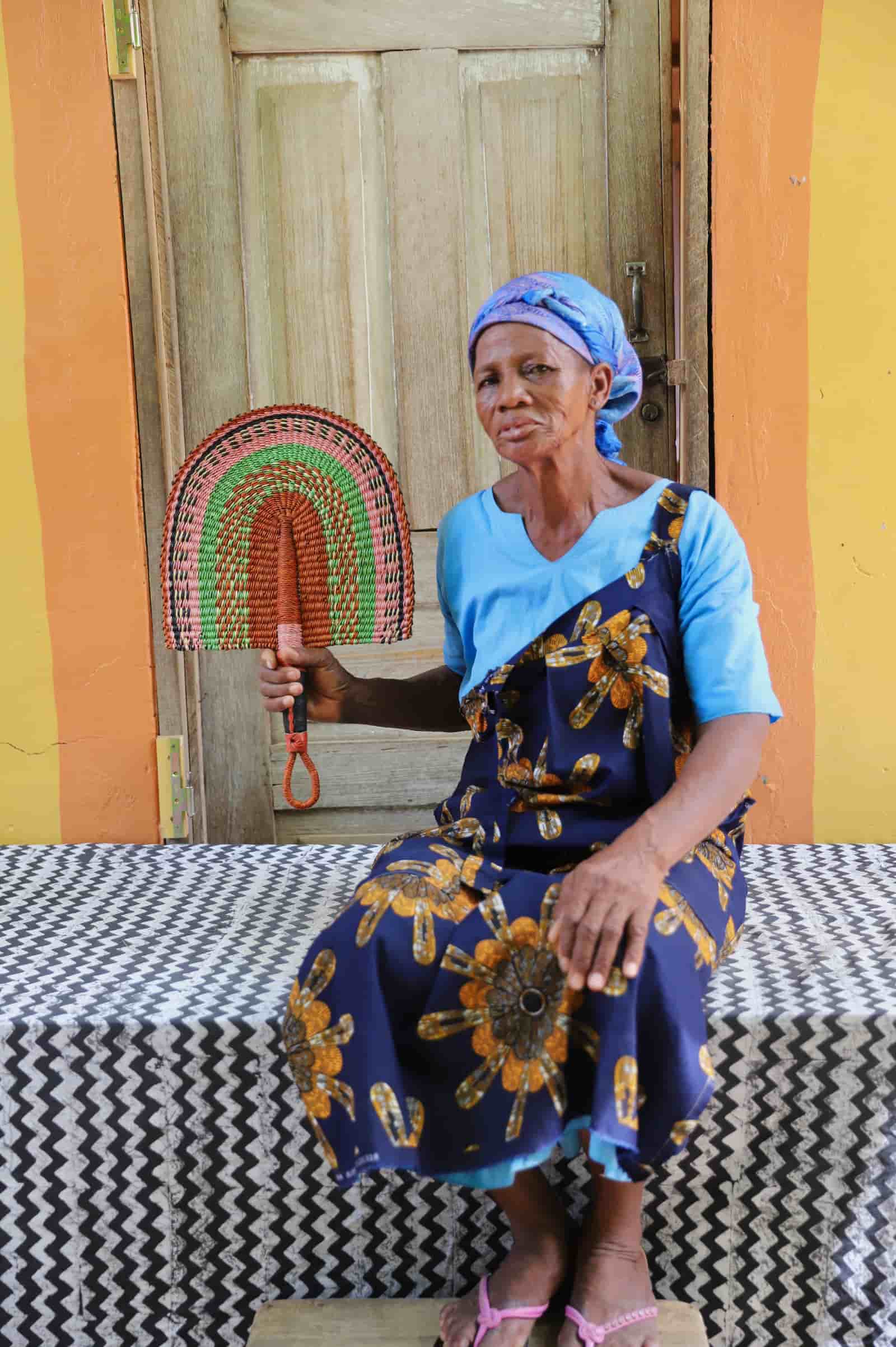 Woman seated holding a Bolga fan in terracotta and green concentric arch pattern, before a wooden door with yellow wall.
