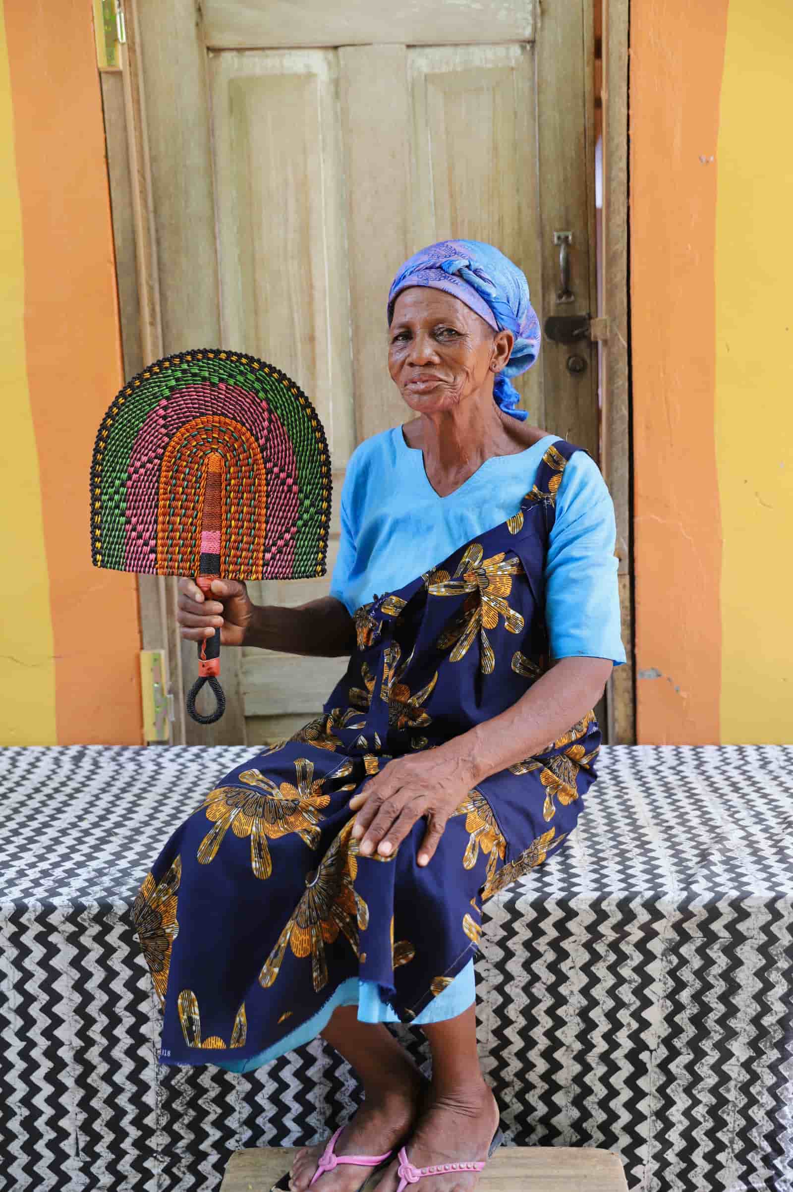 Woman seated holding a Bolga fan with green, pink and amber concentric arch pattern, before a wooden door.
