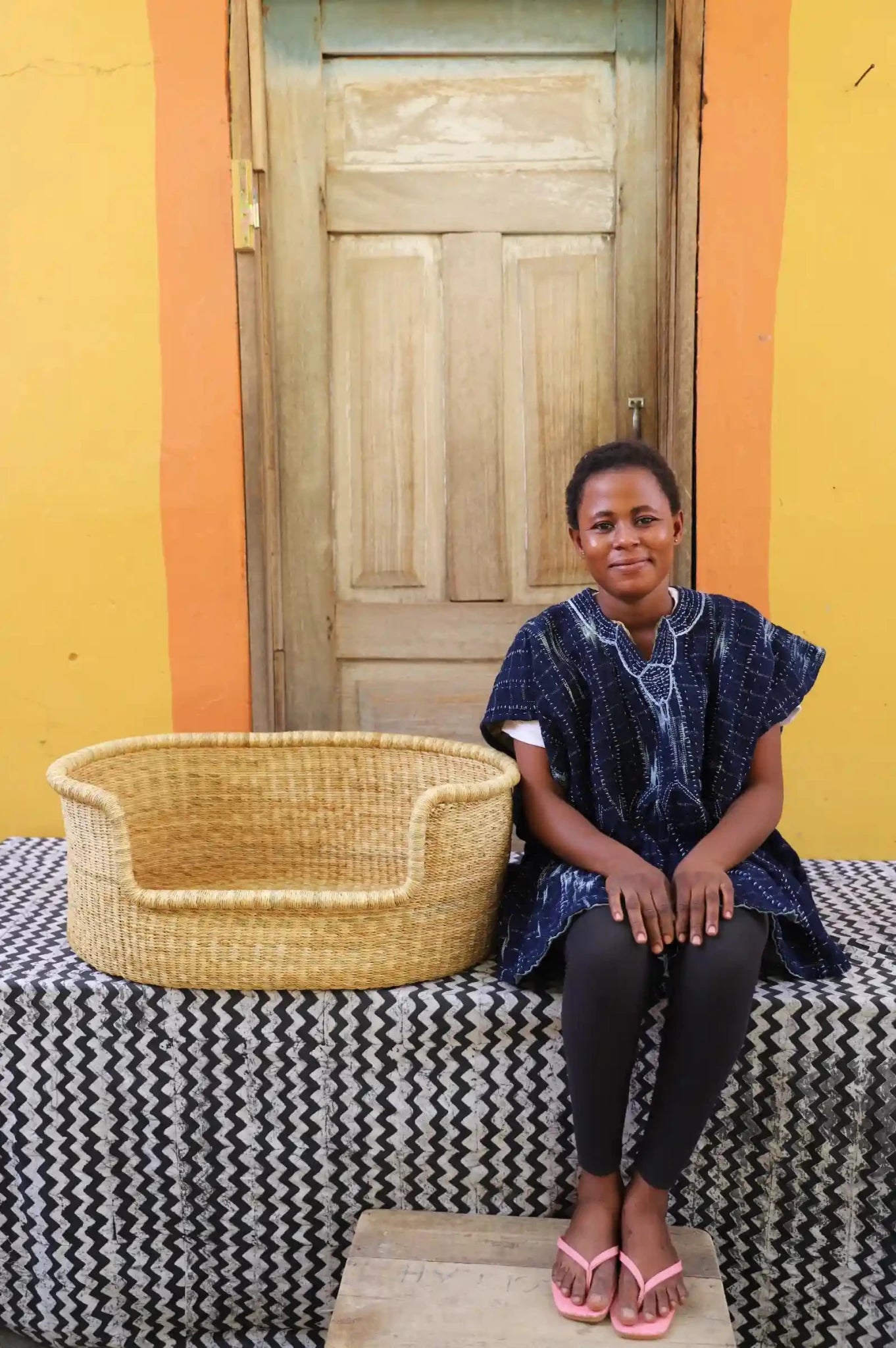Woman sitting on a bench with a woven basket next to a wooden door and yellow wall.

