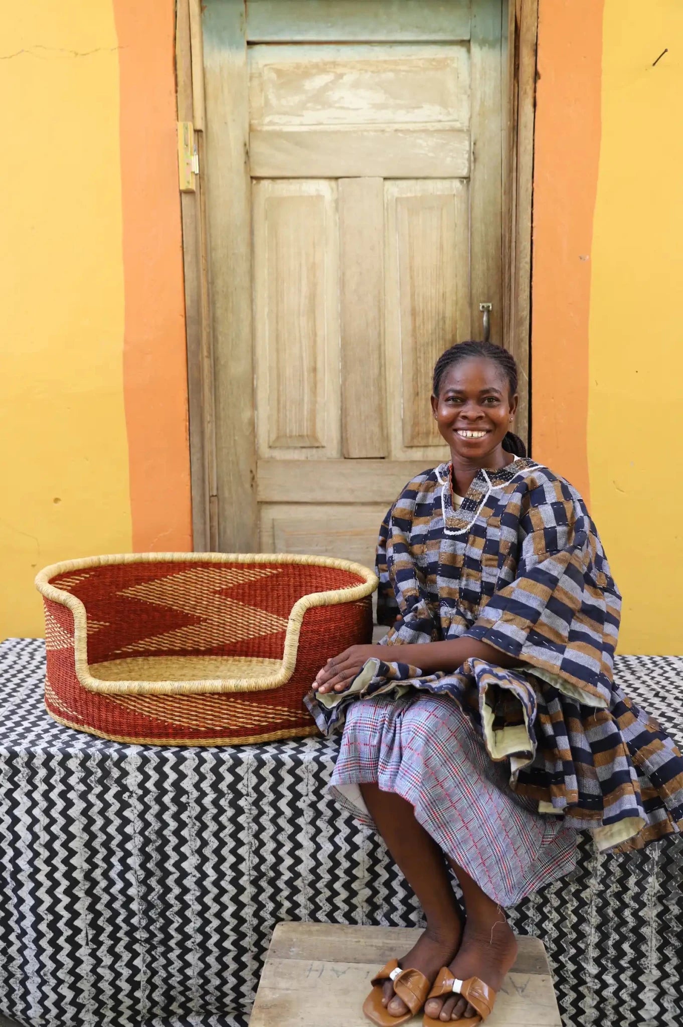 Woman sitting on a bench with a woven basket next to a wooden door and yellow wall.