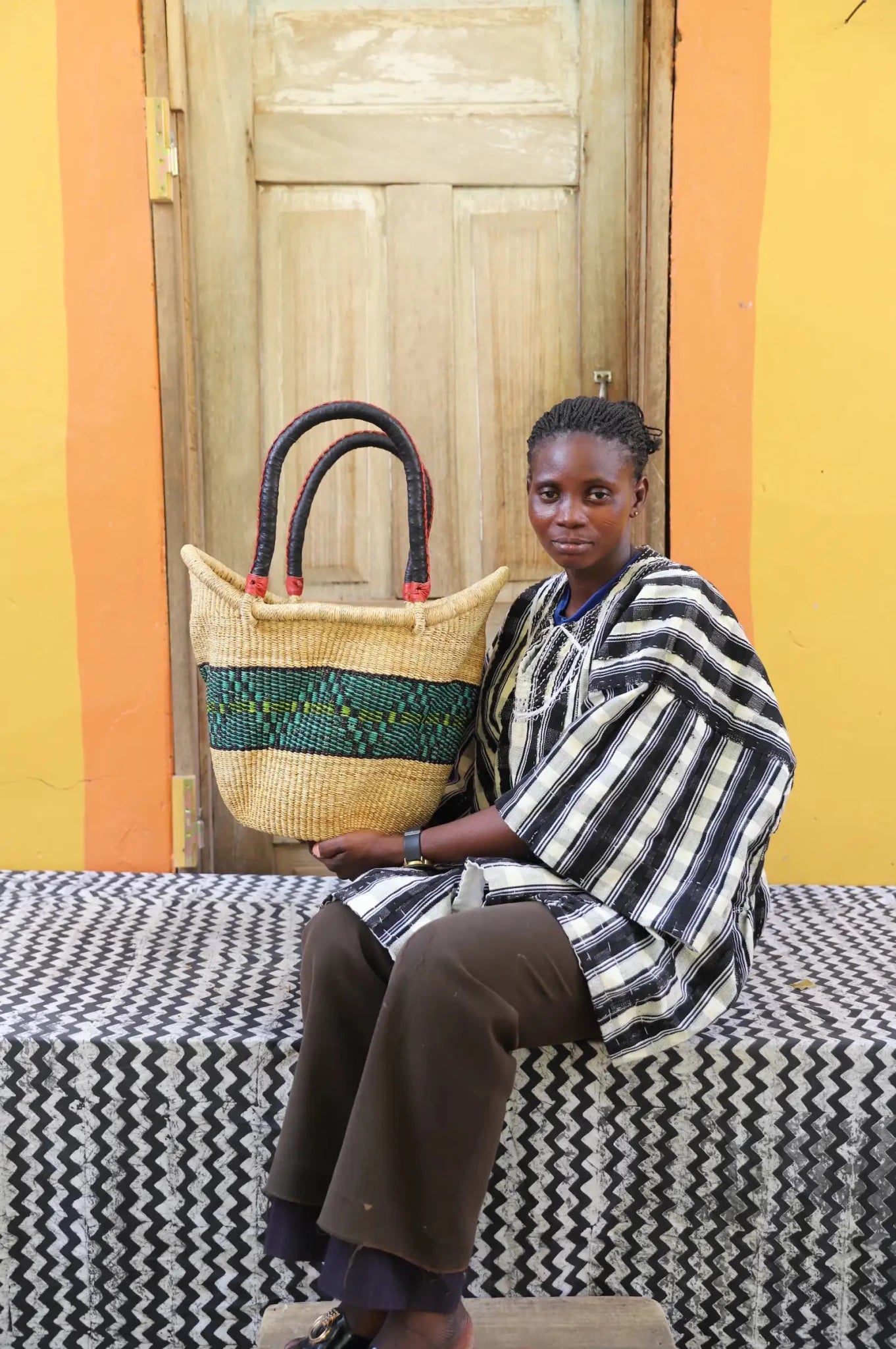 Woman in striped kente smock holding a large Solara Carryall with natural body and green band