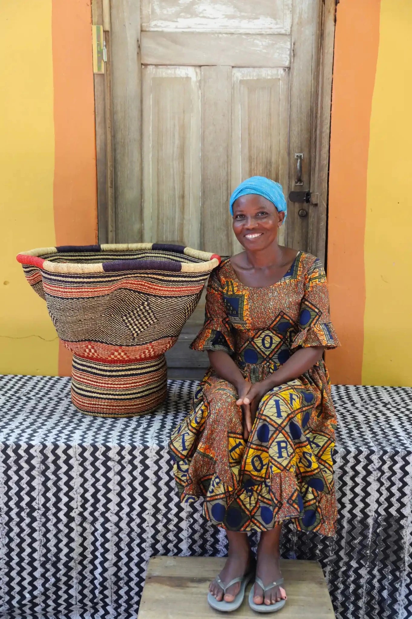 Woman in traditional attire sitting next to a decorative woven basket with stylish straw branching out on a black and white cloth against a wooden door.