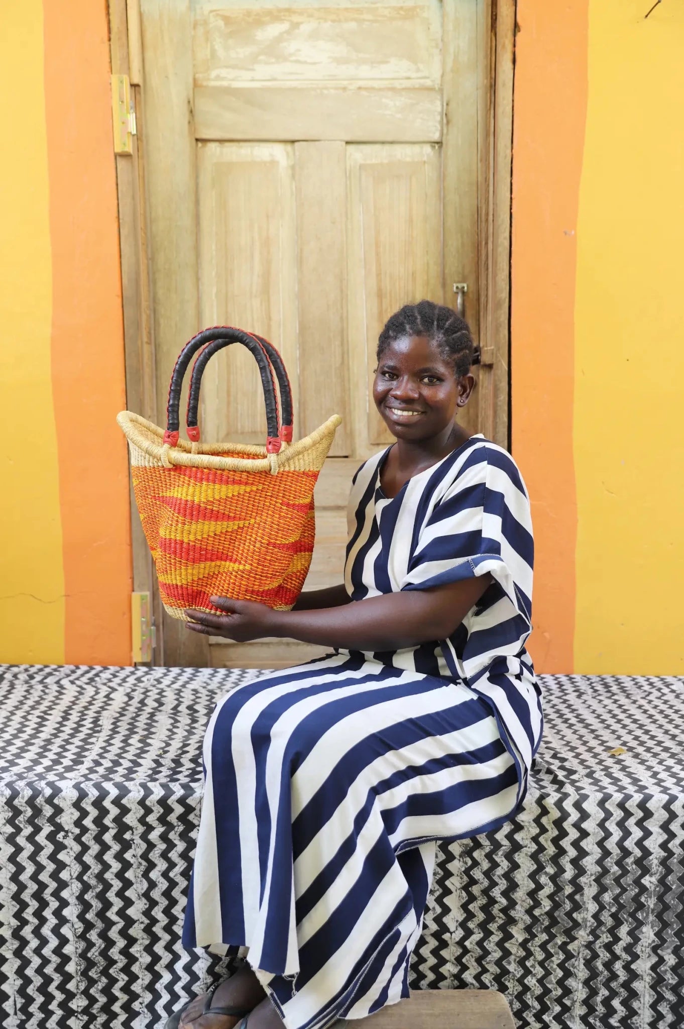 Woman in blue and white striped dress holding a small Solara Carryall with bold orange and yellow diagonal stripe
