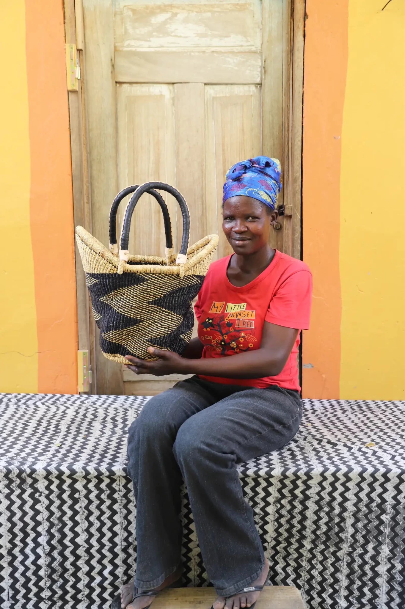 Woman in red top and blue headwrap holding a small Solara Carryall with natural and black zigzag stripe