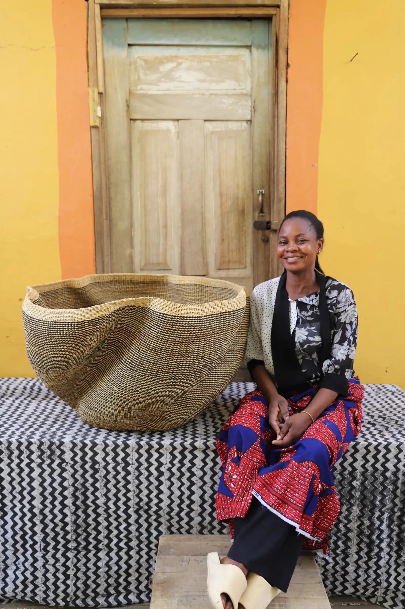 Woman sitting next to a large woven basket in front of a yellow wall with a wooden door.