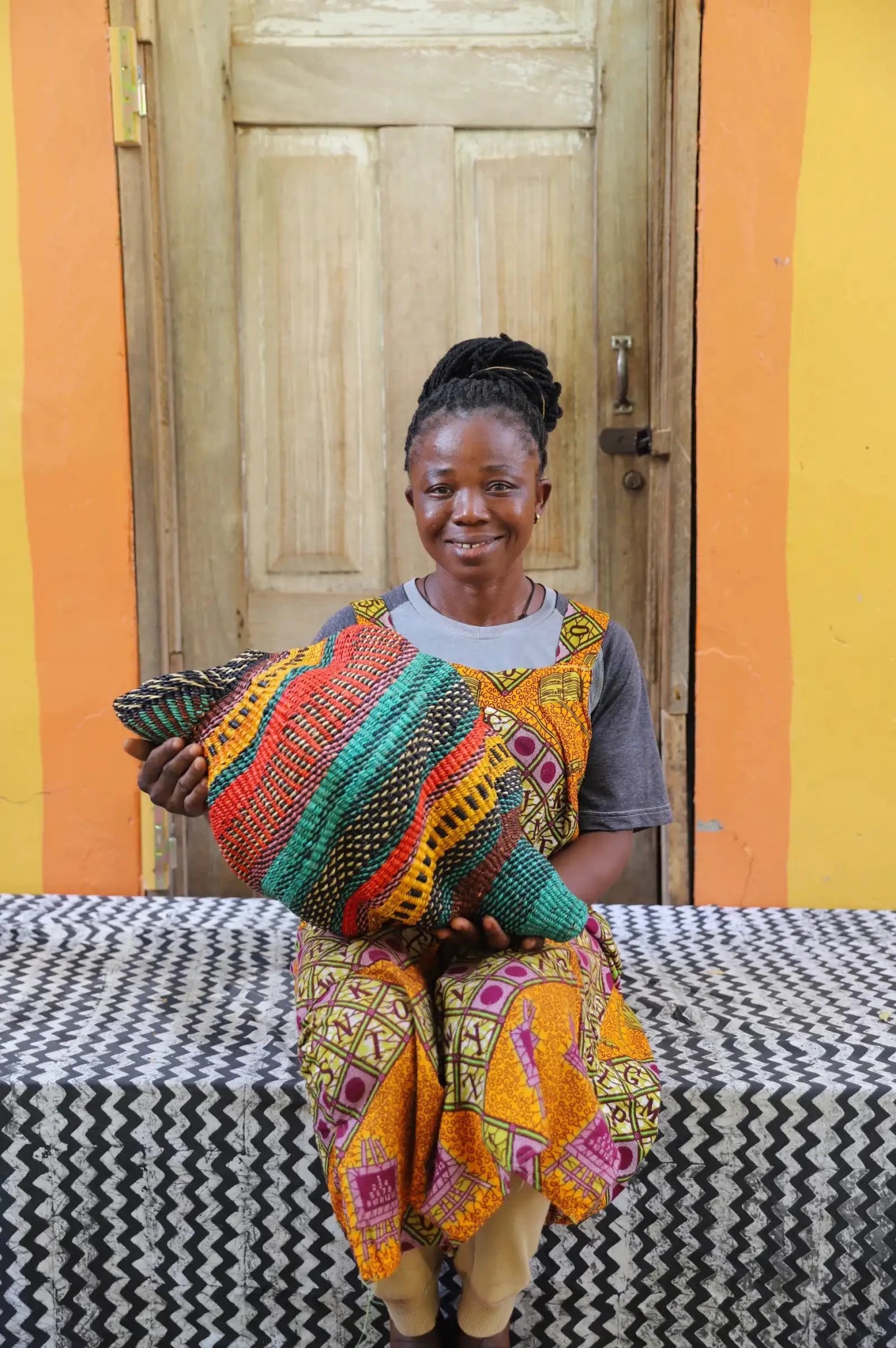 Woman sitting next to a colorful conch-shaped woven piece on a black and white cloth against a wooden door.