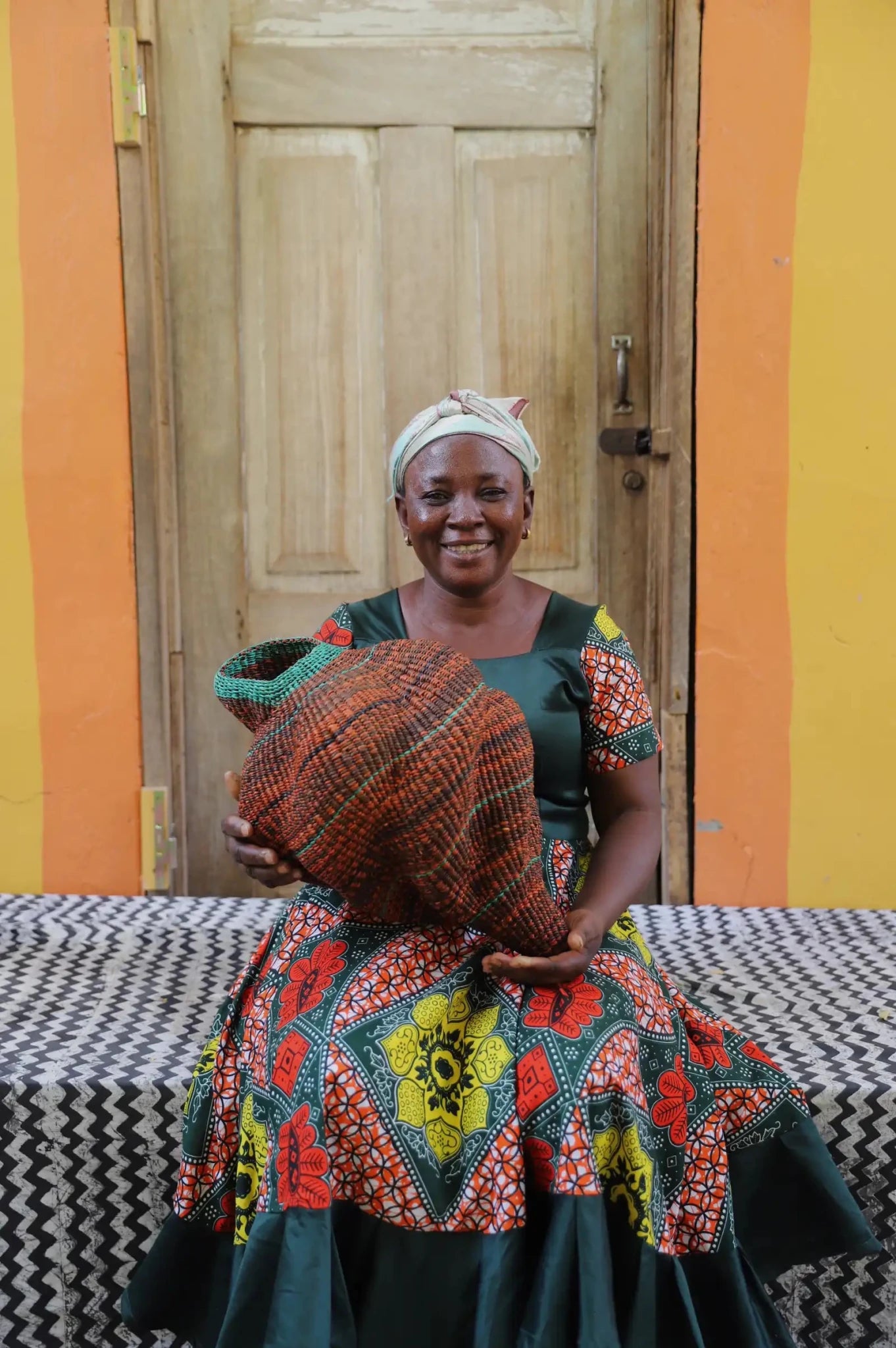 Woman sitting next to a colorful conch-shaped woven piece on a black and white cloth against a wooden door.