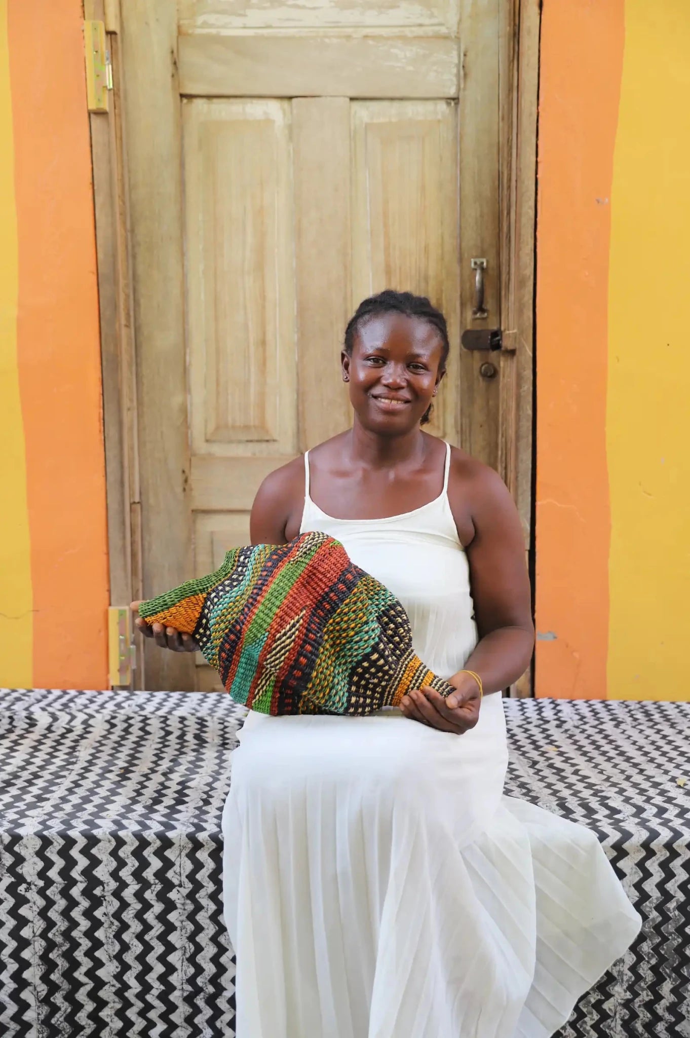 Woman sitting next to a colorful conch-shaped woven piece on a black and white cloth against a wooden door.