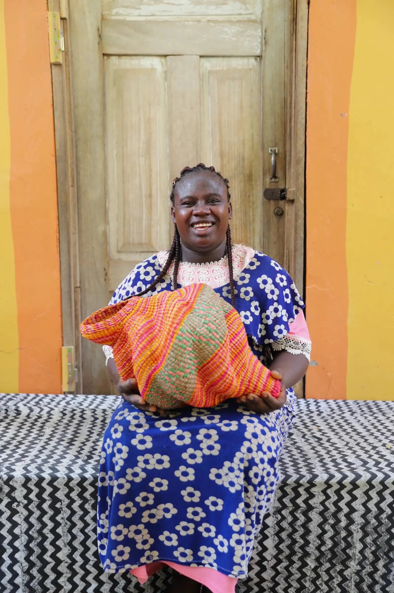 Woman sitting next to a colorful conch-shaped woven piece on a black and white cloth against a wooden door.