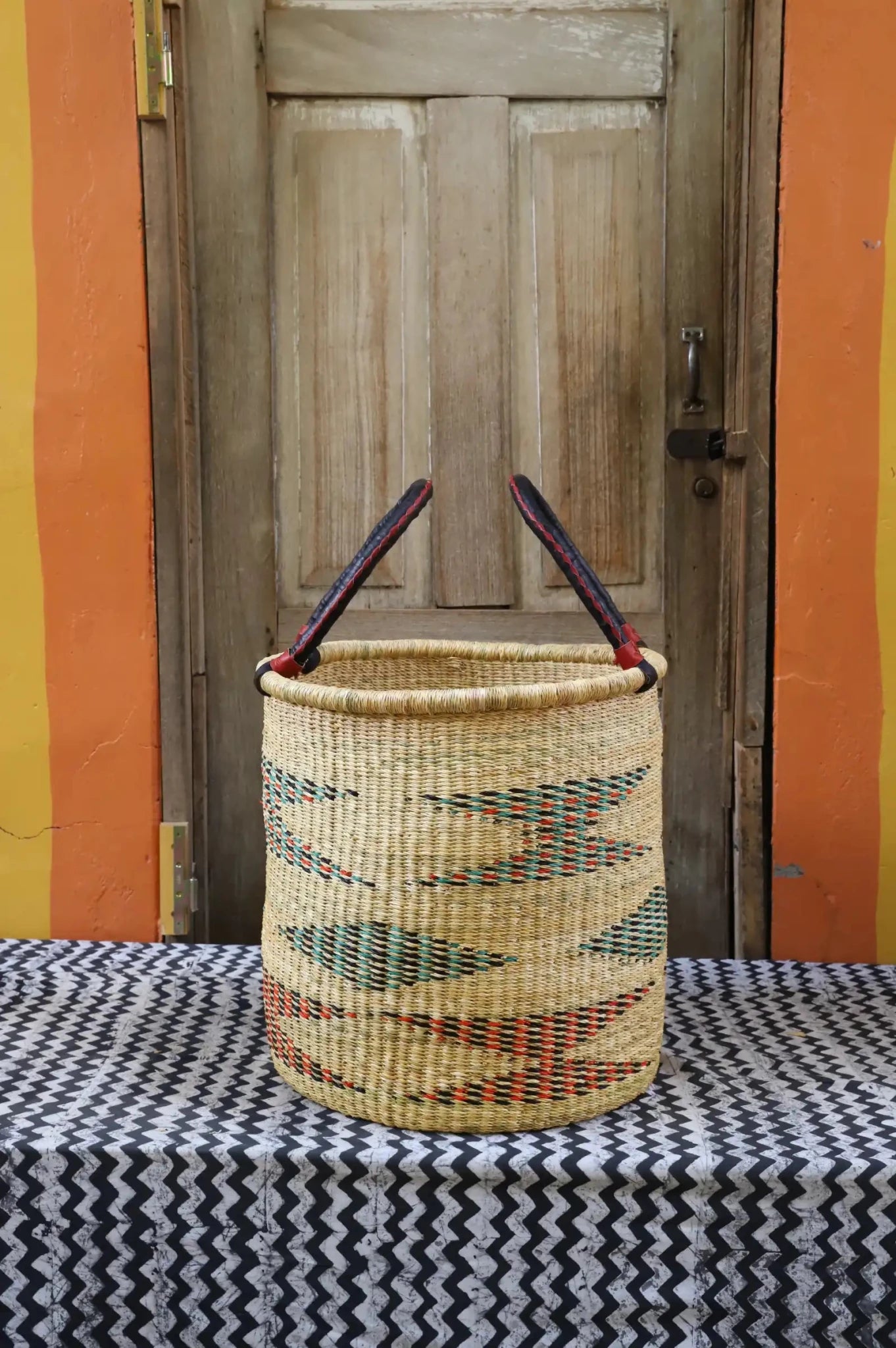 Colourful hand woven laundry basket sitting on black and white patterned cloth in front of a wooden door