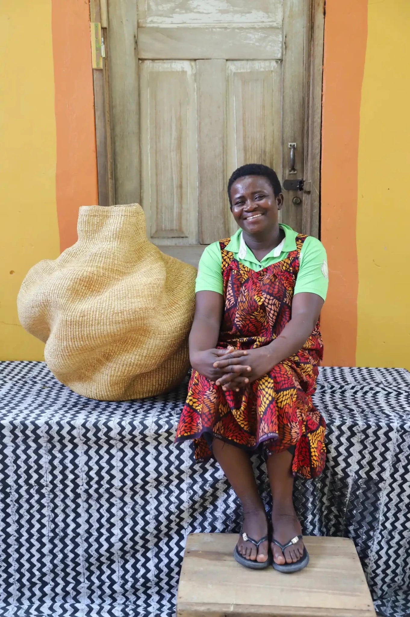 An artisan seated next to a handcrafted decorative basket  on a black and white cloth against a wooden door.