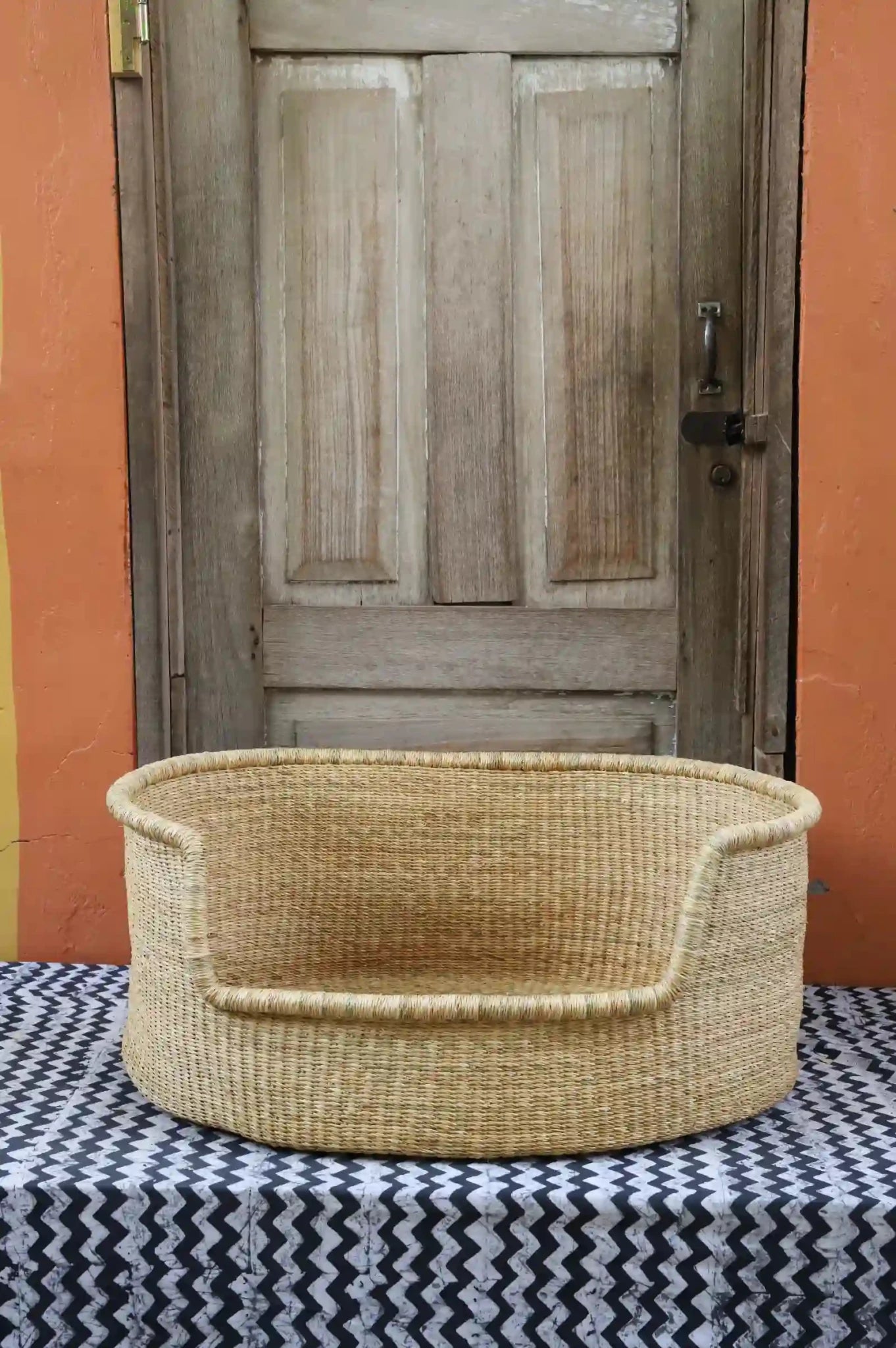 Woven pet bed with geometric pattern on a textured surface in front of a wooden door.