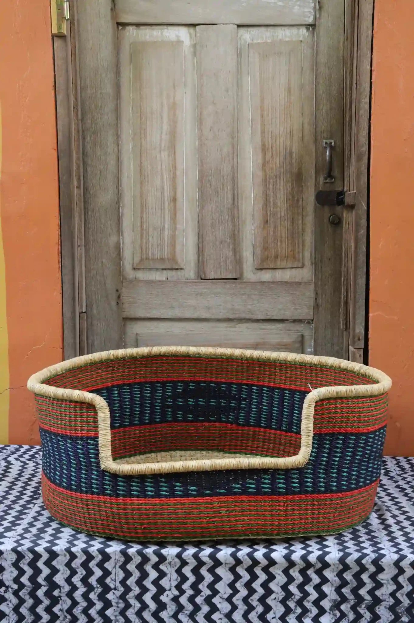 Woven pet bed with geometric pattern on a textured surface in front of a wooden door.