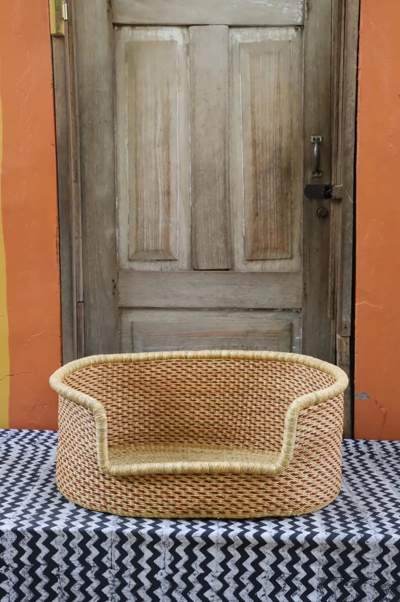 Woven pet bed with geometric pattern on a textured surface in front of a wooden door.