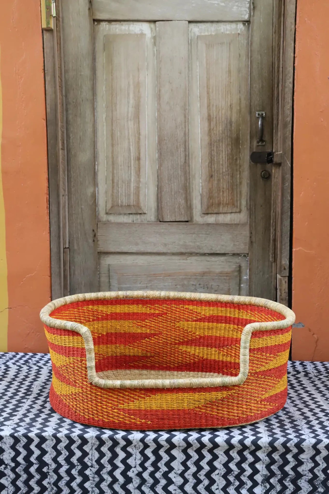 Woven pet bed with geometric pattern on a textured surface in front of a wooden door.