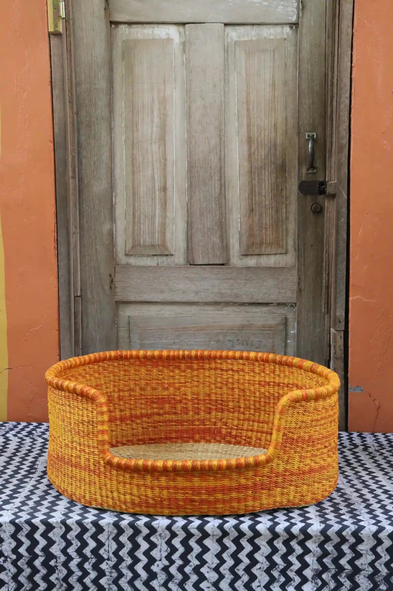Woven pet bed with geometric pattern on a textured surface in front of a wooden door.