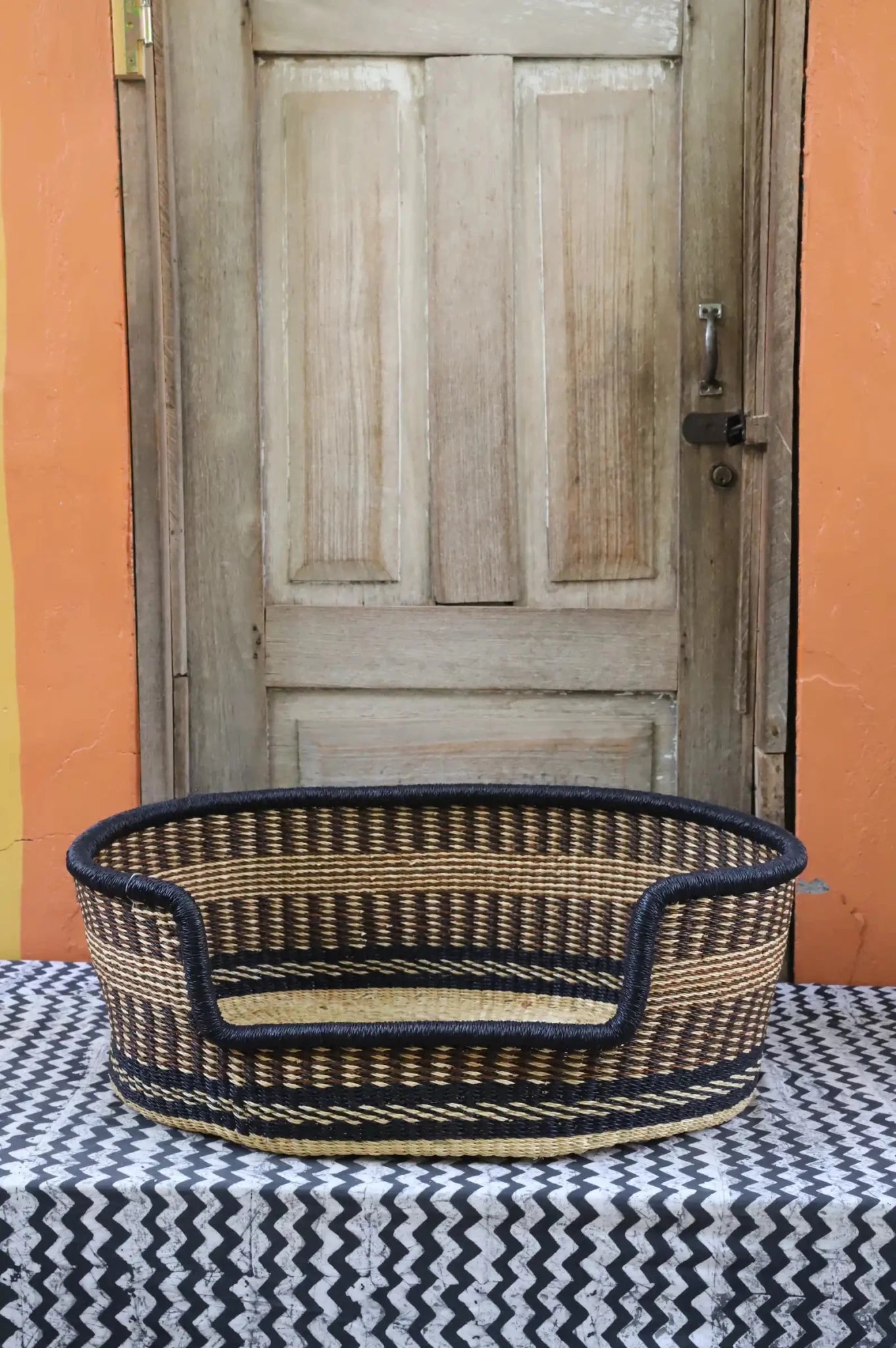 Woven pet bed with geometric pattern on a textured surface in front of a wooden door.