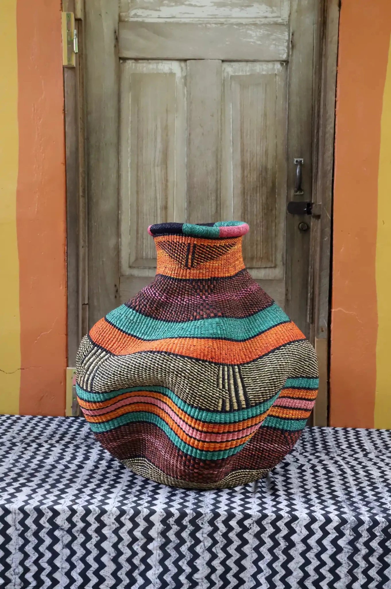 Colorful woven piece on a black and white patterned surface with a wooden door in the background.