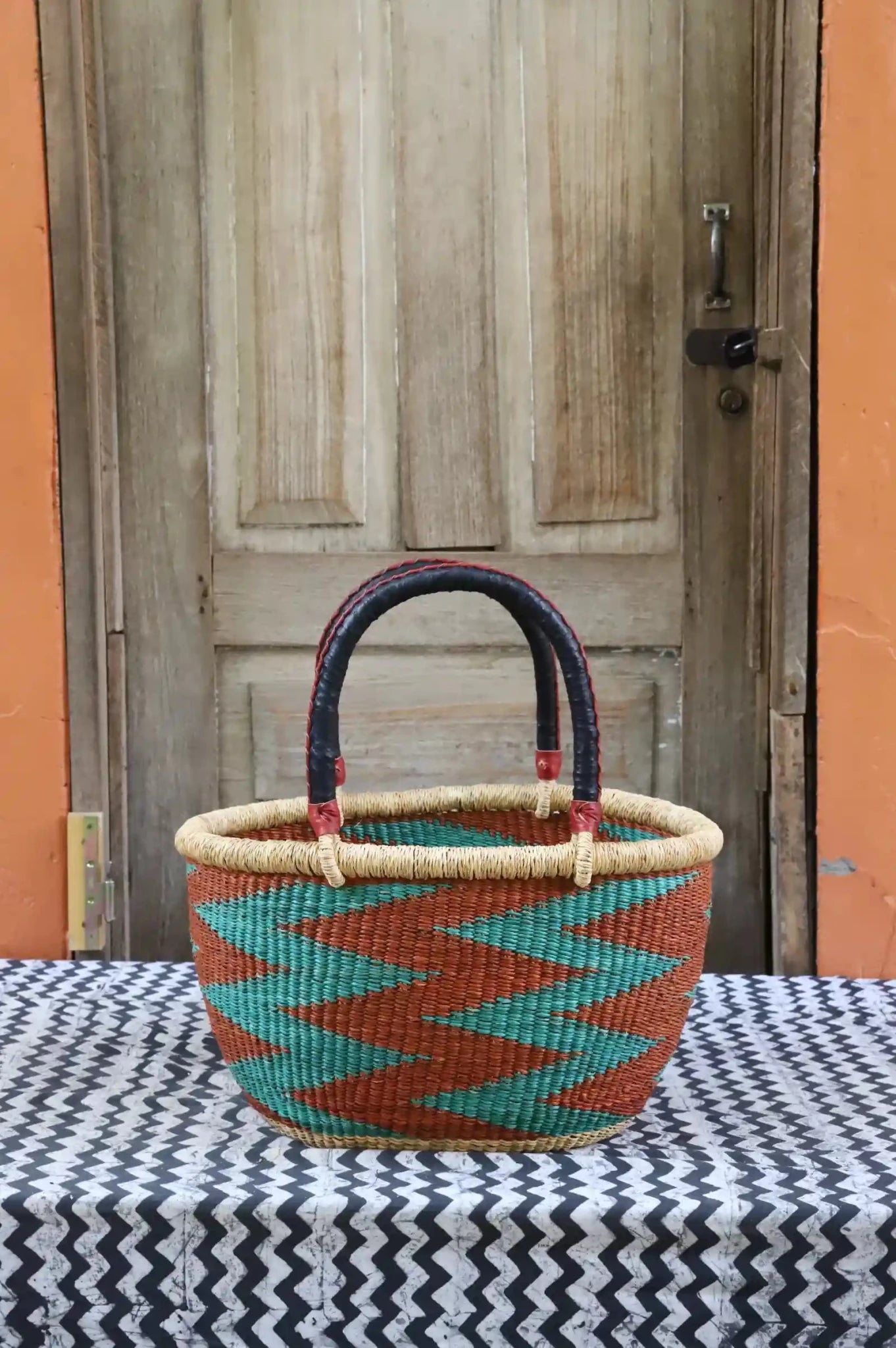 Woven carry on bag with premium leather handles on a black and white cloth against a wooden door.