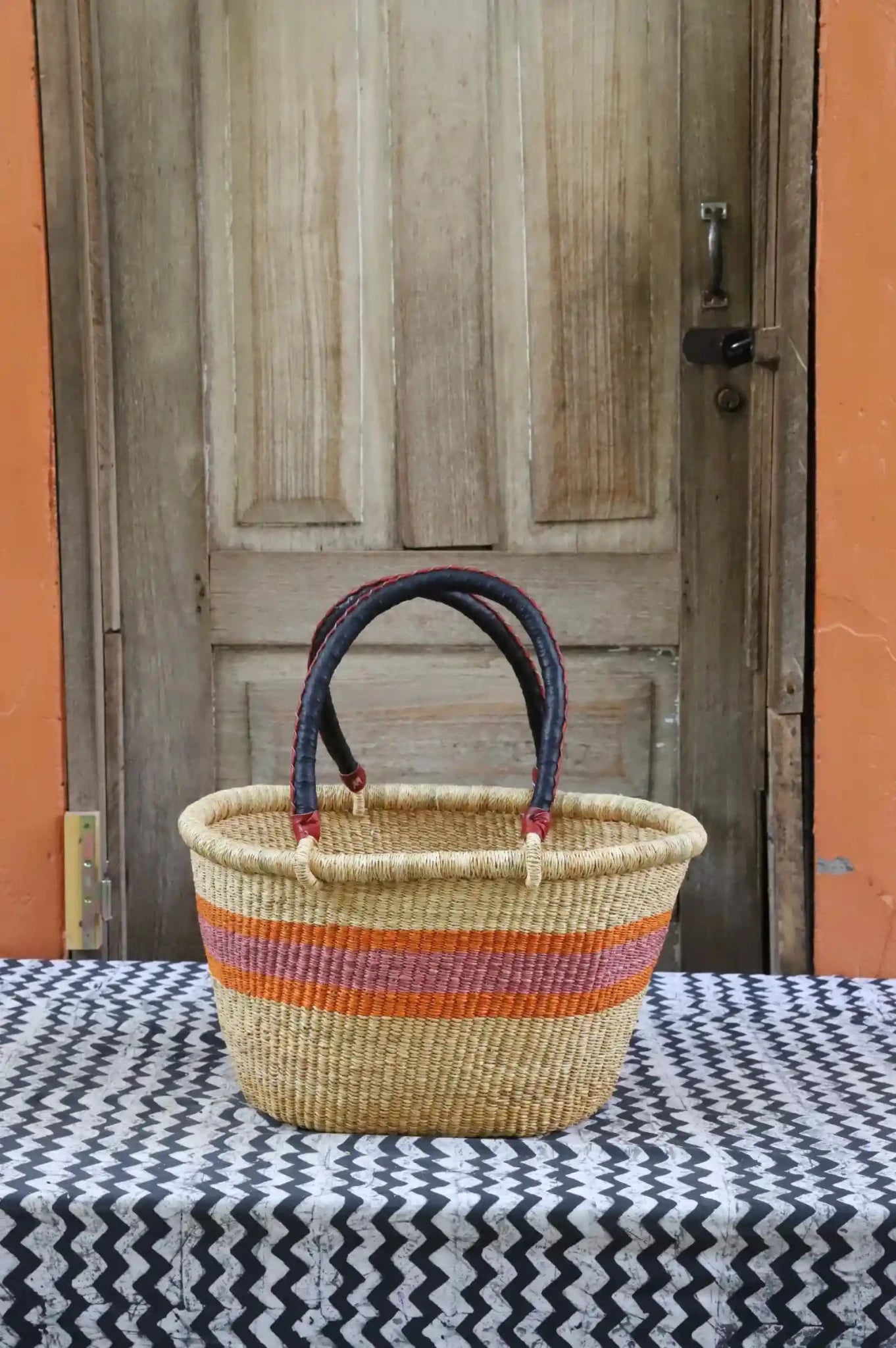 Woven carry on bag with premium leather handles on a black and white cloth against a wooden door.
