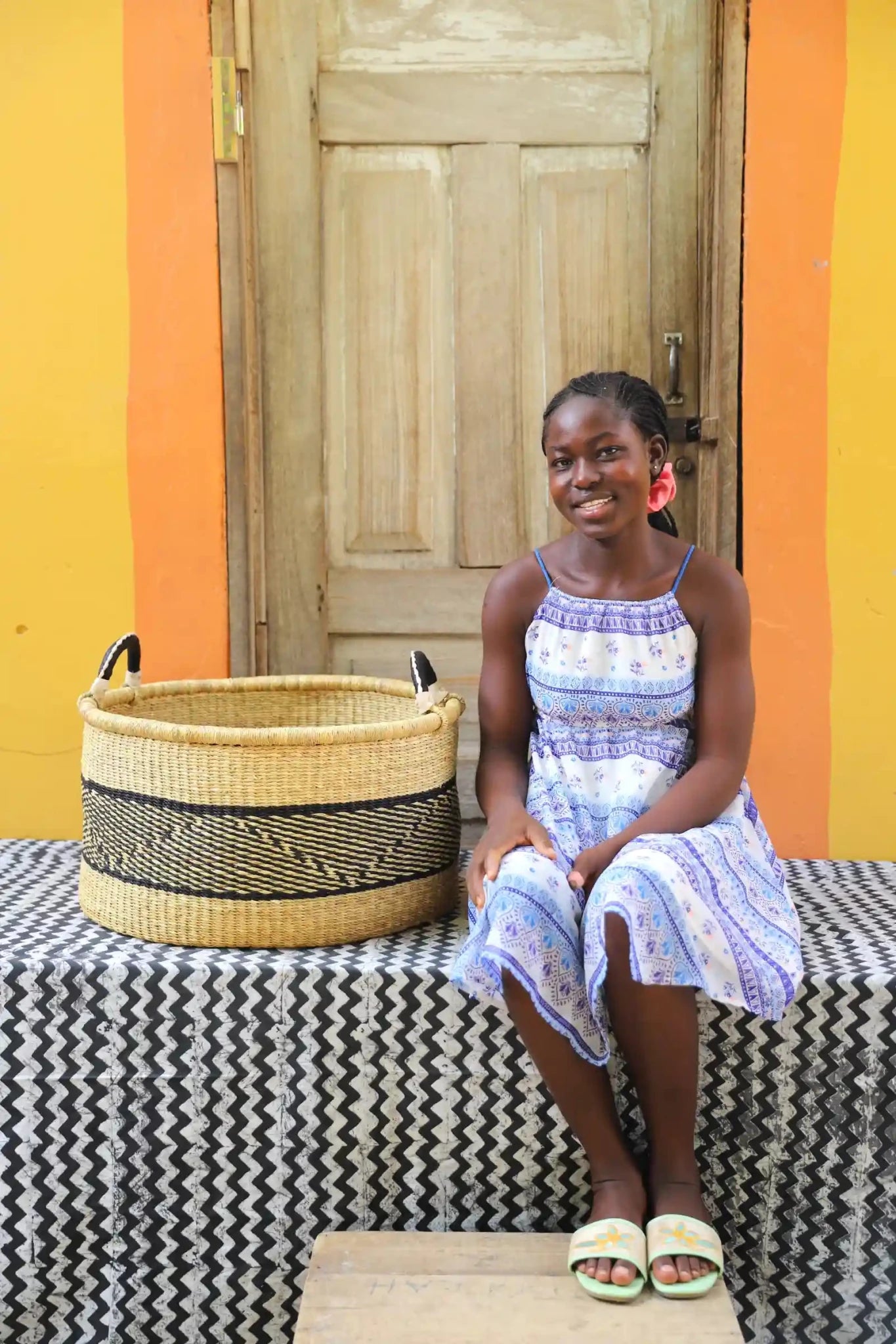 Woven Tub (Large) with woman weaver handwoven in Ghana sitting on black and white cloth and wooden door in background
