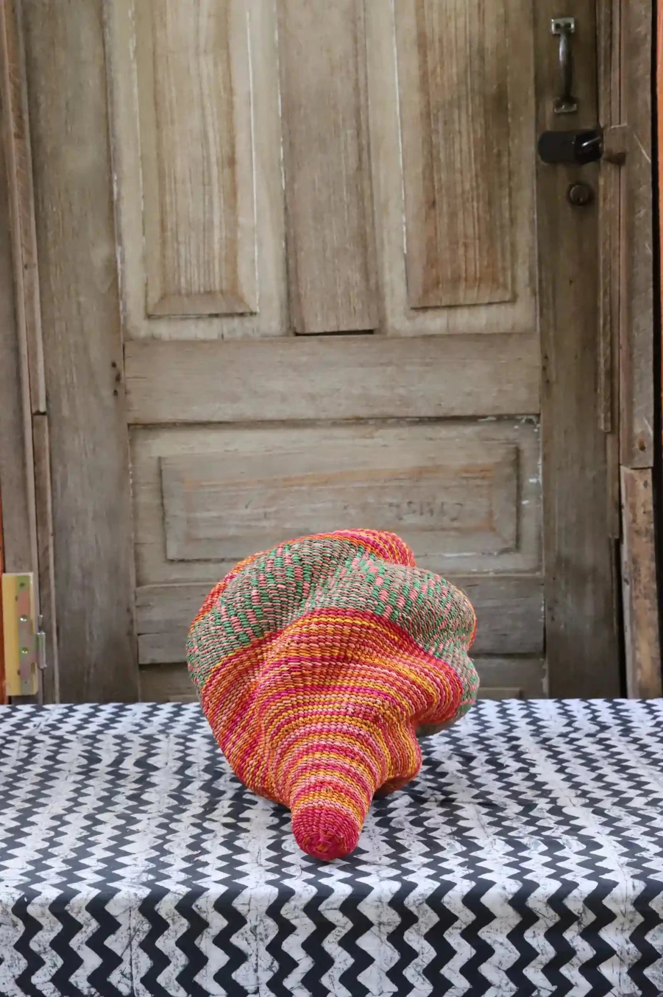 Colorful conch-shaped woven piece on a black and white cloth against a wooden door.