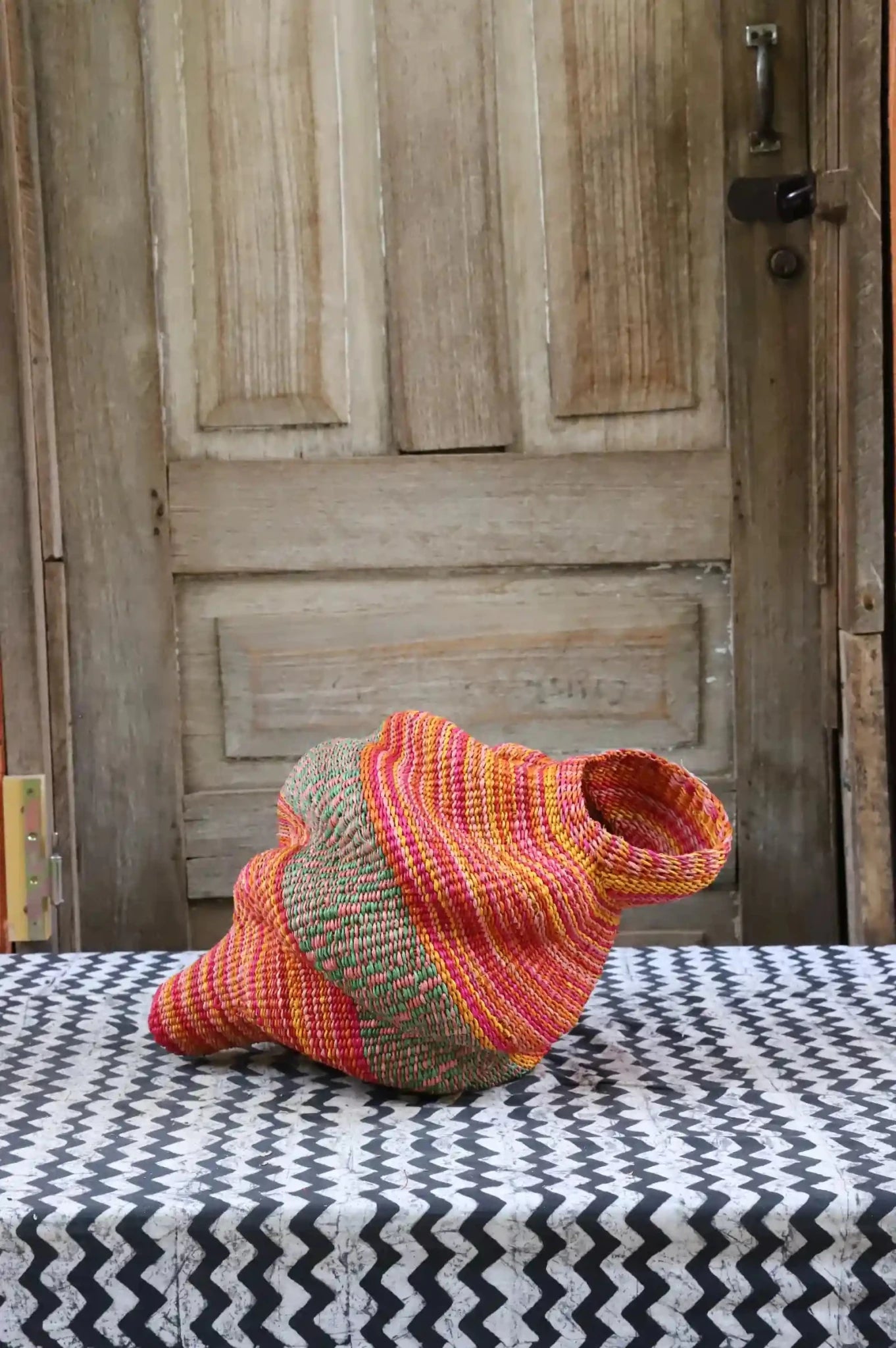 Handwoven Katayeli-tia art basket in pink, green and amber flowing stripes, on a black and white chevron cloth.