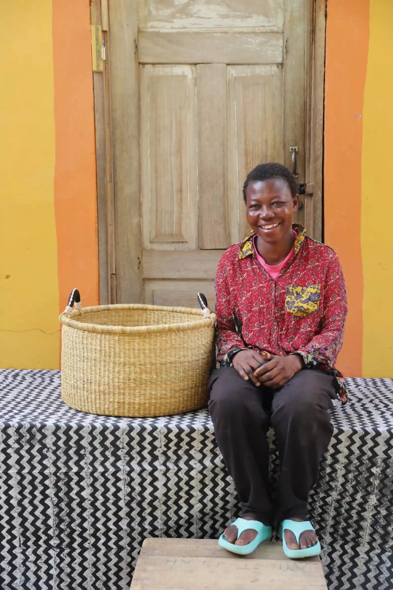 Woven Tub (Medium) with woman weaver handwoven in Ghana sitting on black and white cloth and wooden door in background

