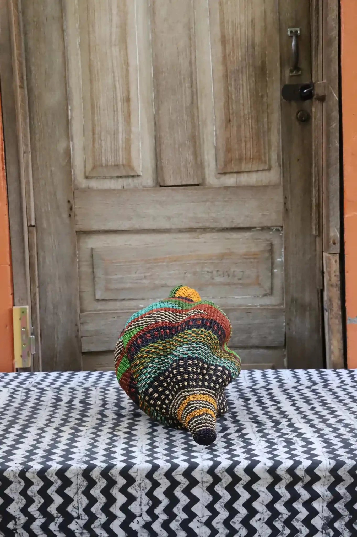 Colorful conch-shaped woven piece on a black and white cloth against a wooden door.