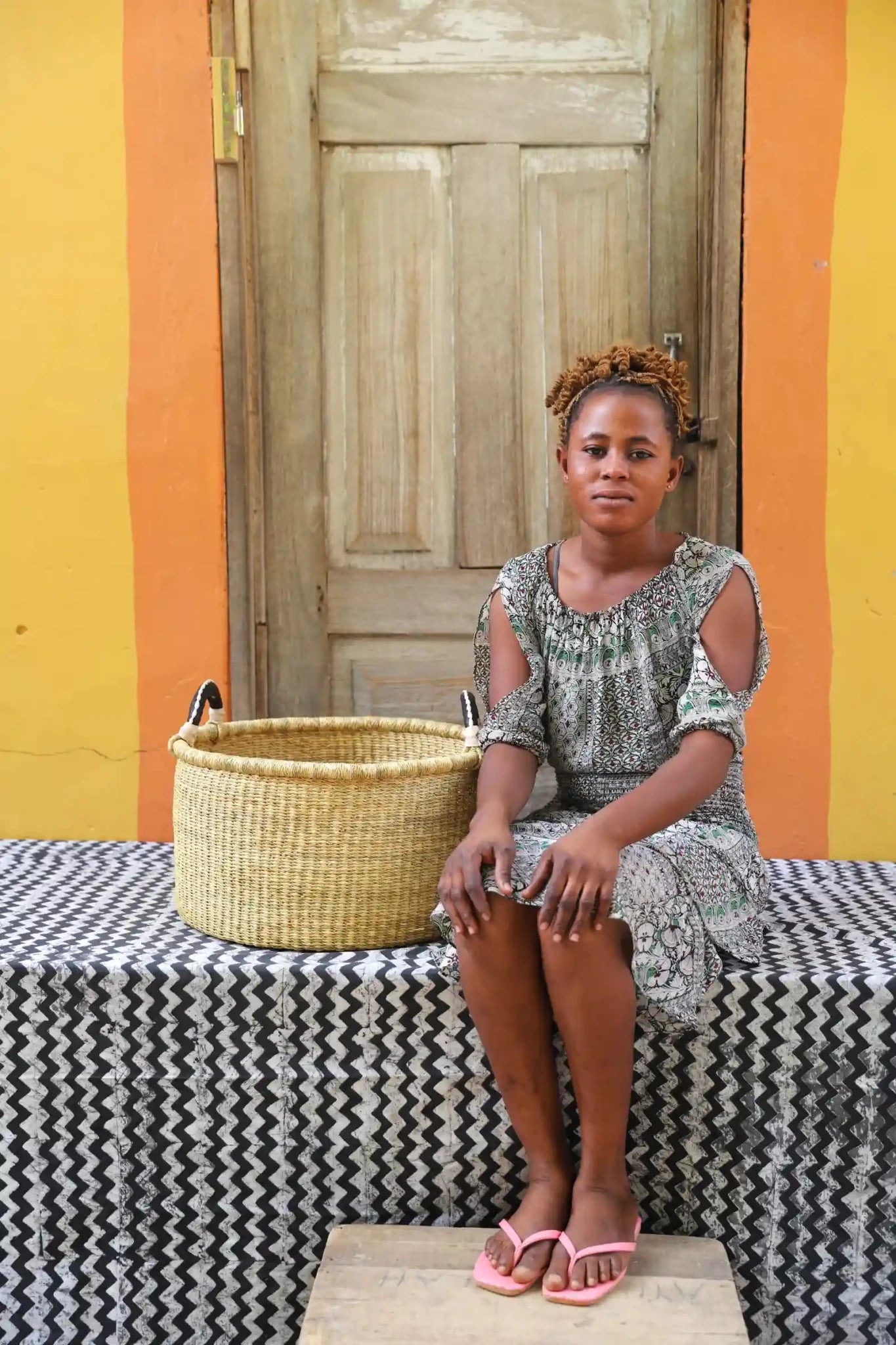 Woven Tub (Small) with woman weaver handwoven in Ghana sitting on black and white cloth and wooden door in background
