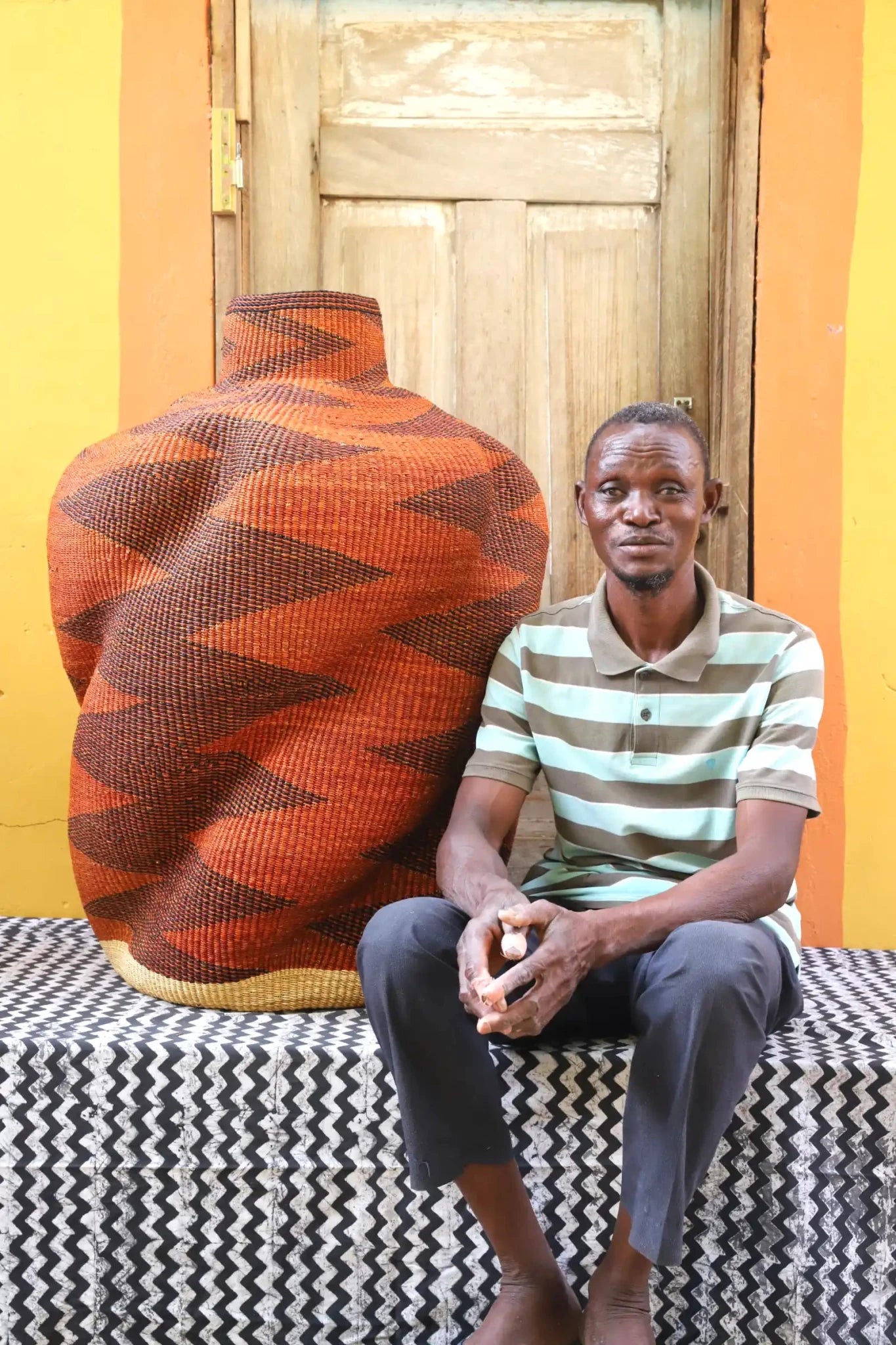 Man sitting next to a curvy, handwoven décor piece on a black and white cloth against a wooden door.