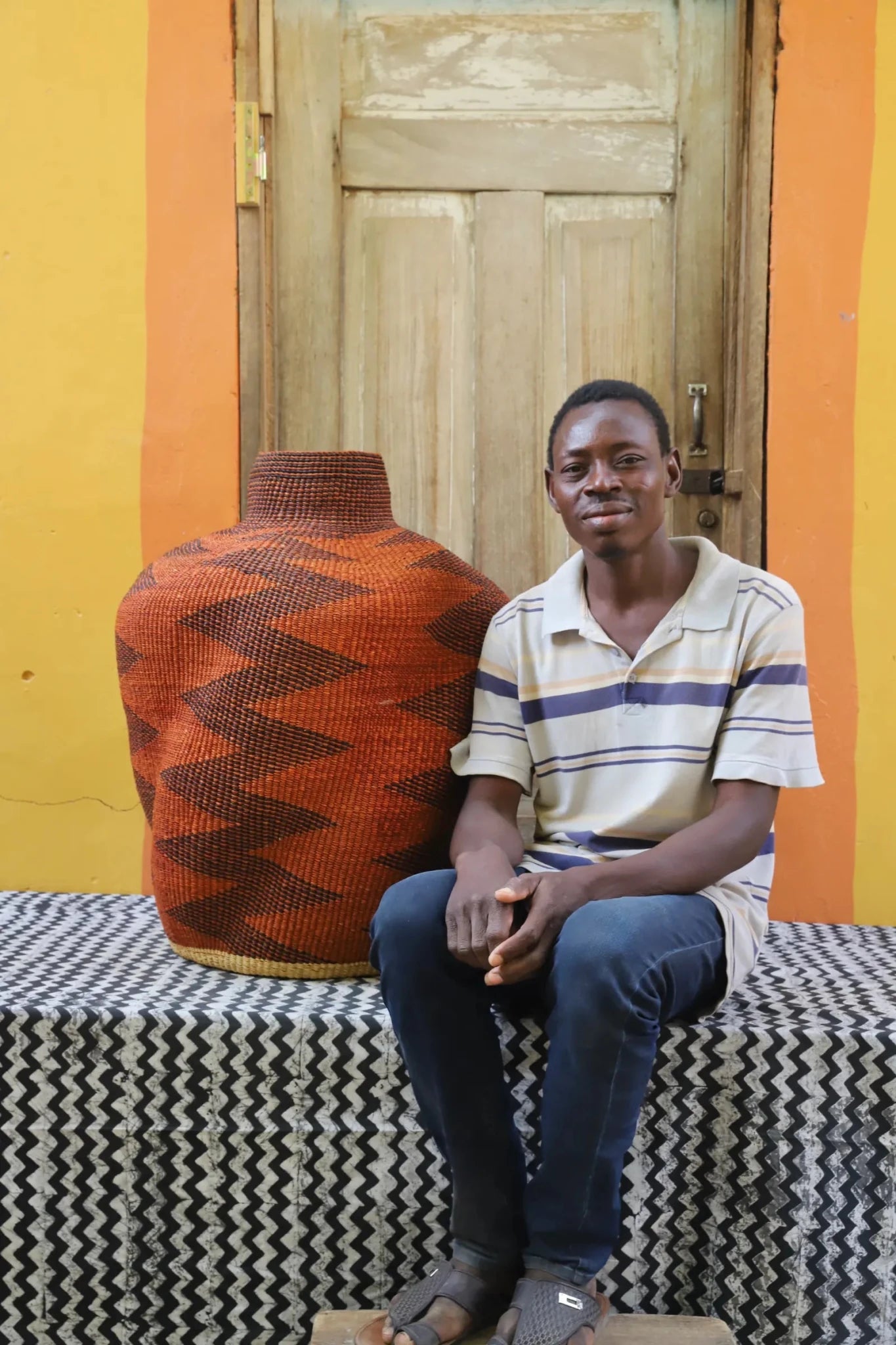 Man sitting with a curvy, handwoven décor piece on a black and white cloth against a wooden door.