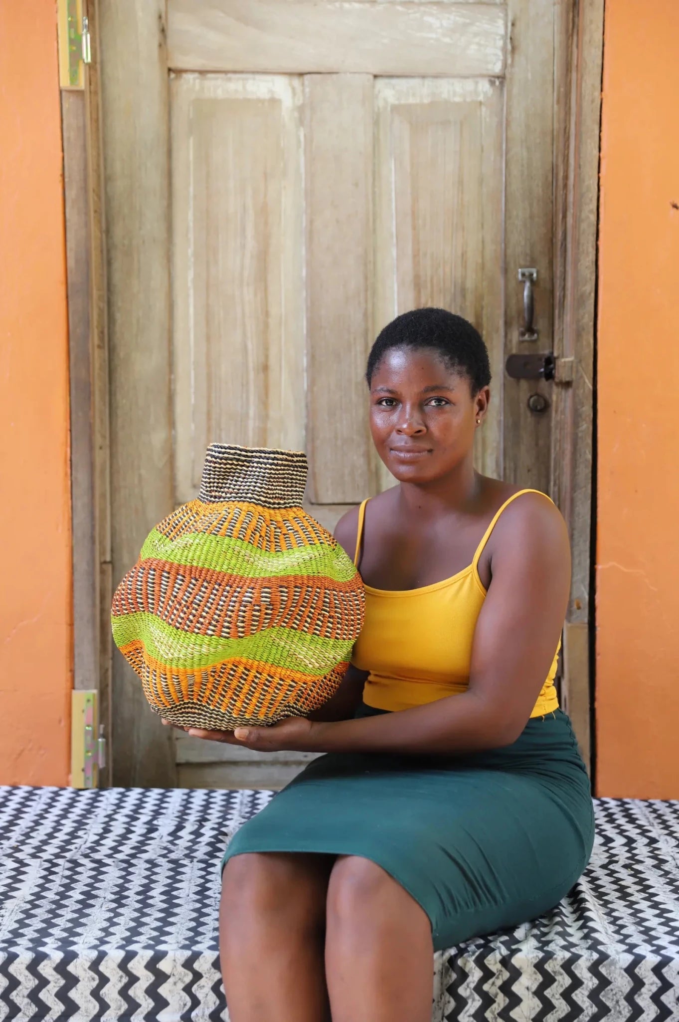 An artisan seated next to a handcrafted decorative basket  on a black and white cloth against a wooden door.