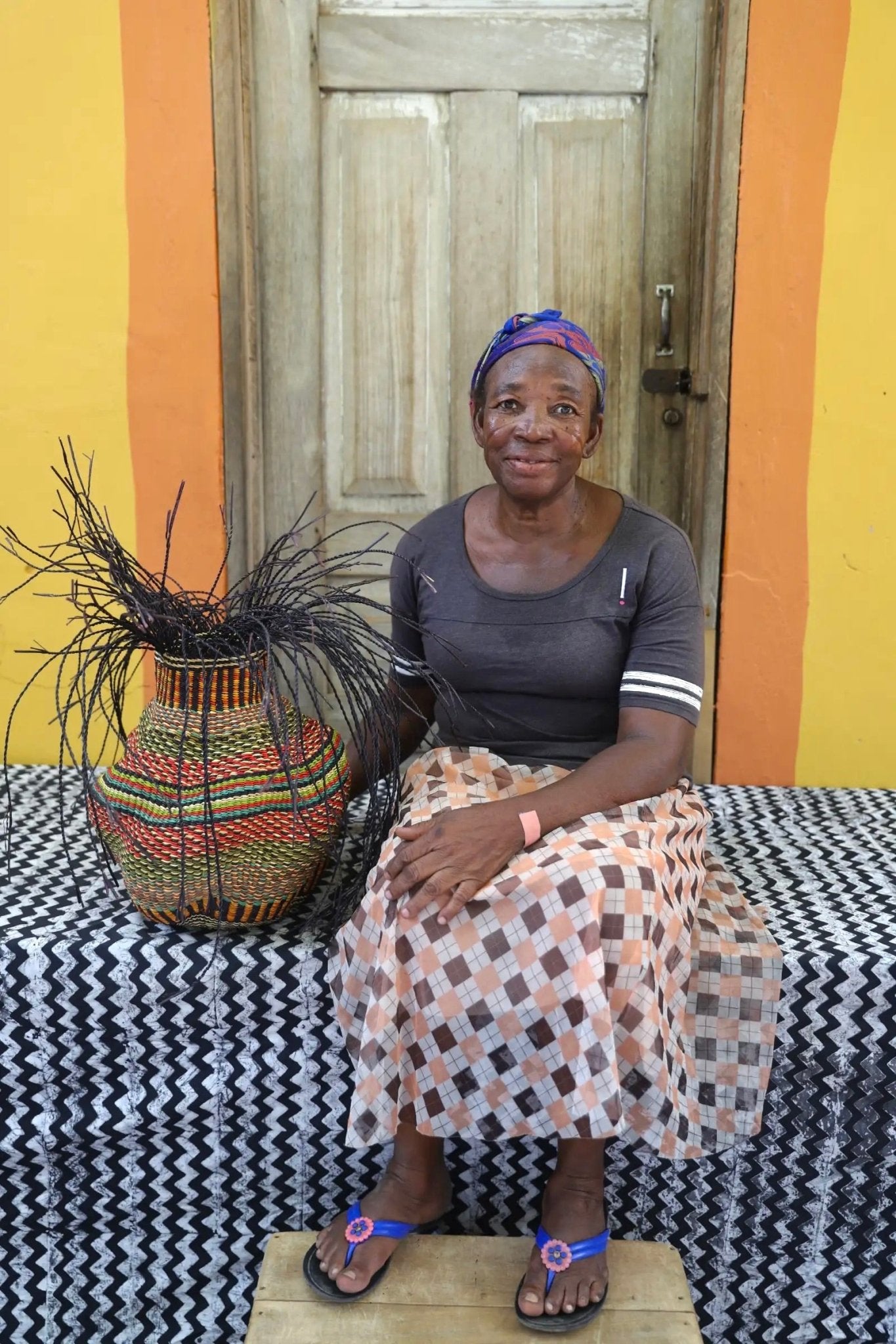 A woman sitting next to a colorful, handwoven piece sitting on a black and white cloth with decorative straw branching out. 