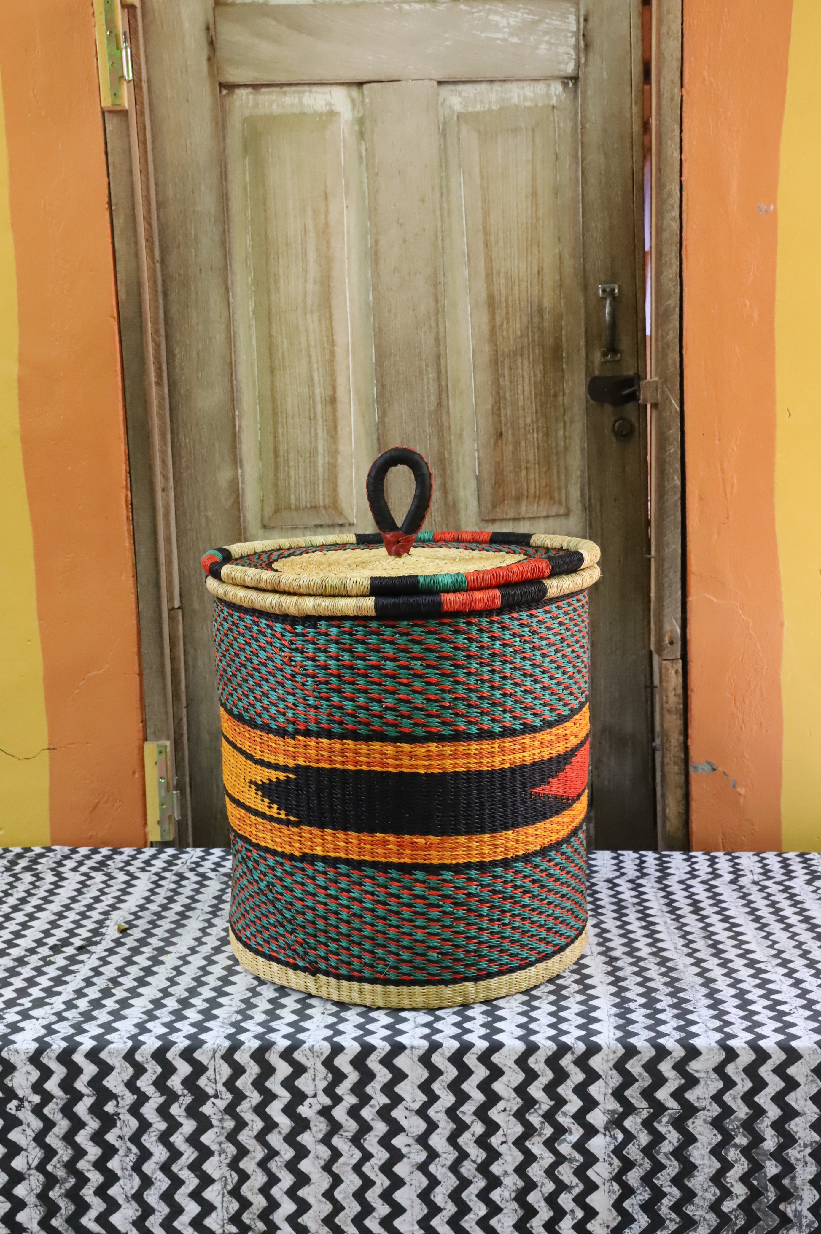 Large coiled-rim lidded laundry basket in dark weave with orange and black banding, styled on a chevron cloth.