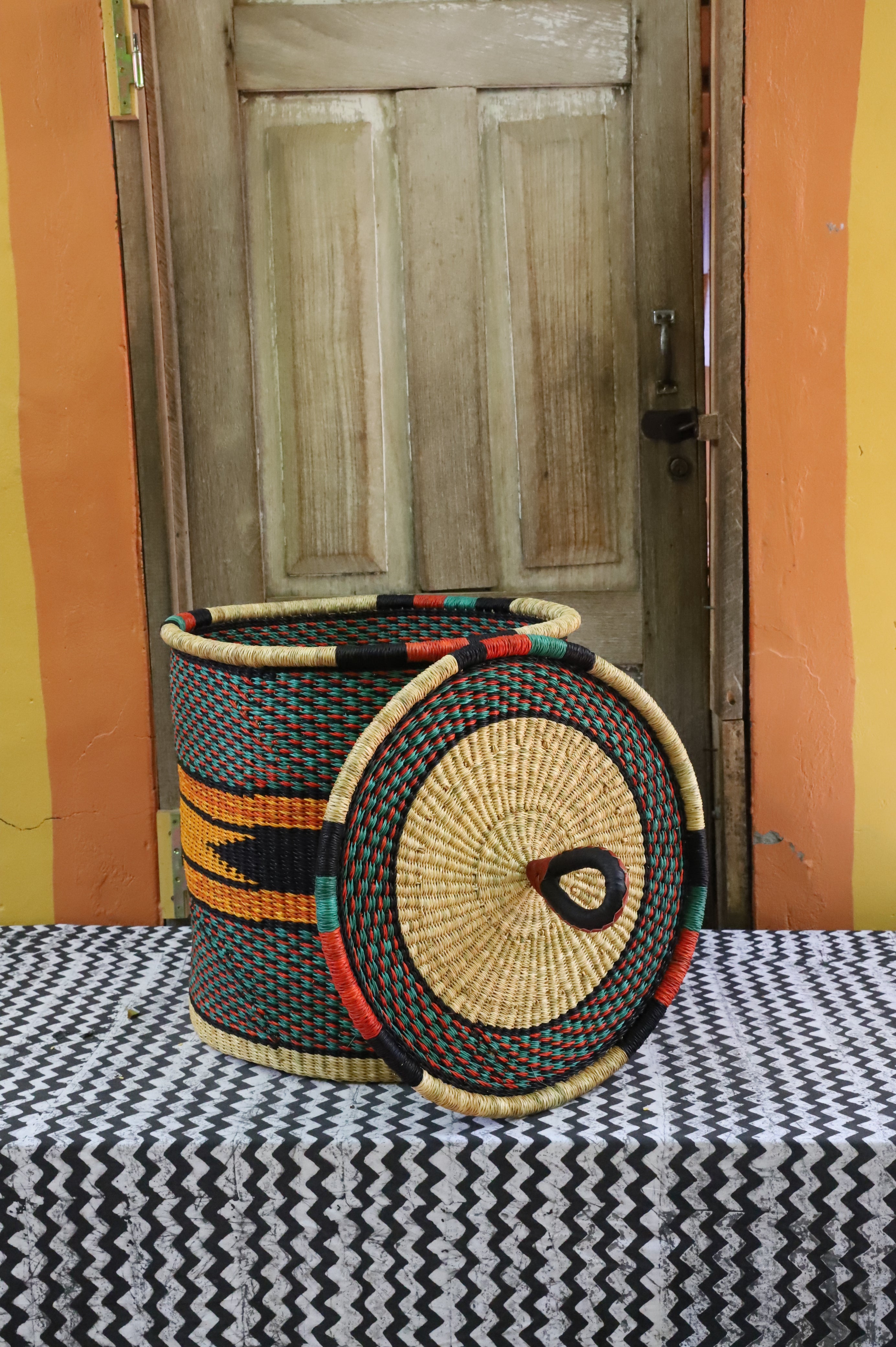 Large coiled-rim laundry basket in dark weave with orange and black banding, lid resting alongside on a chevron cloth.