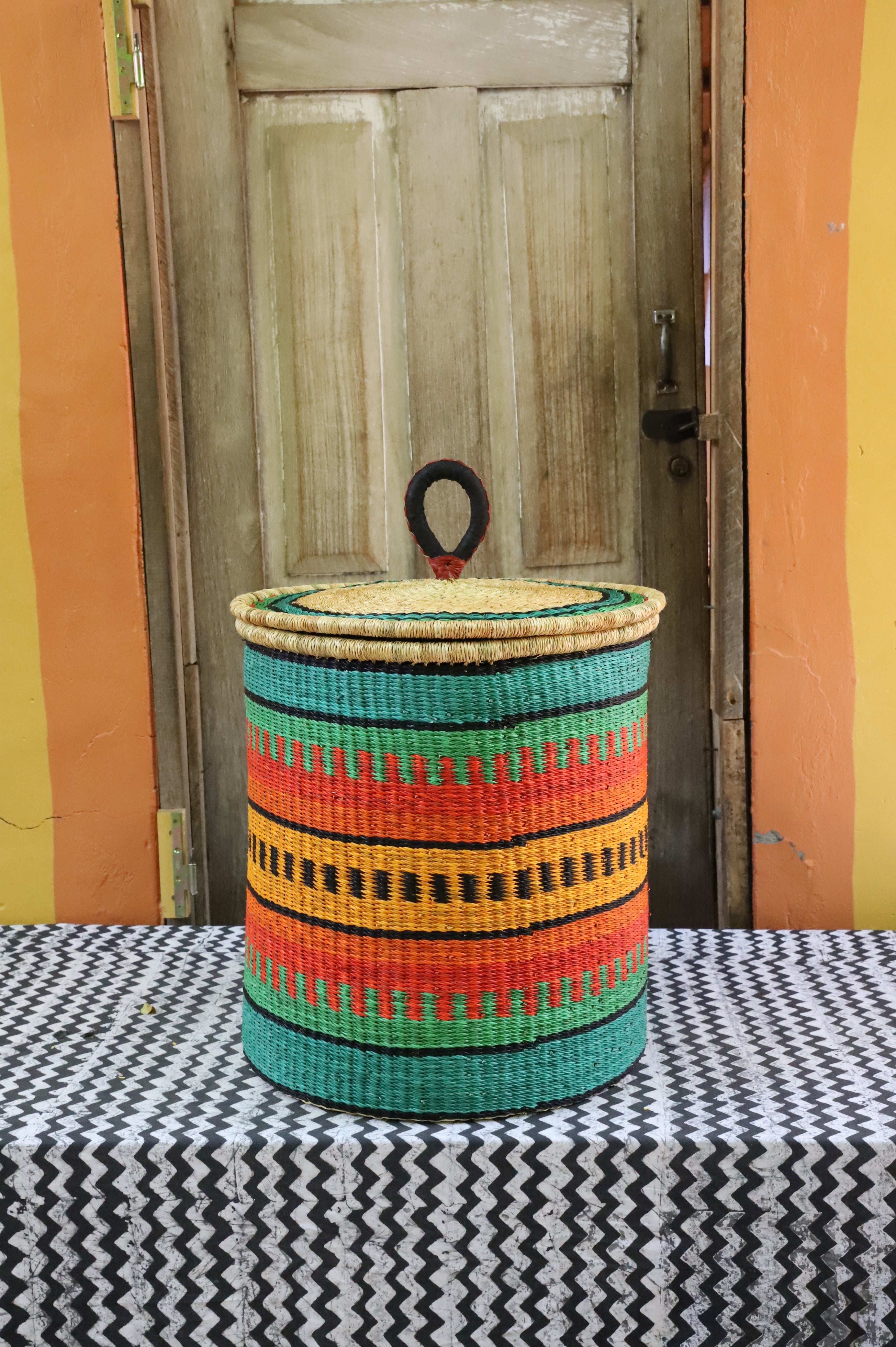 Large coiled-rim lidded laundry basket in green, orange and black striped weave, styled on a chevron cloth.